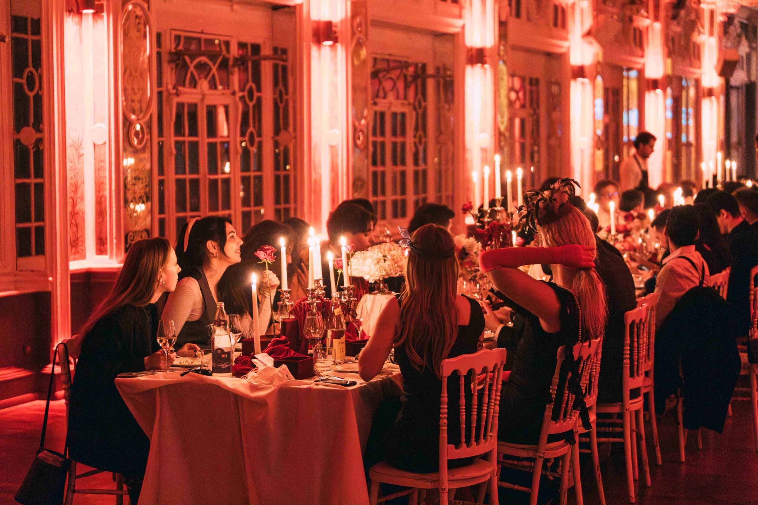 A formal dinner party with guests seated at a long table in an elegant, warmly lit room with tall windows and ornate decor. Candles and floral arrangements adorn the table, creating a cozy and festive atmosphere.