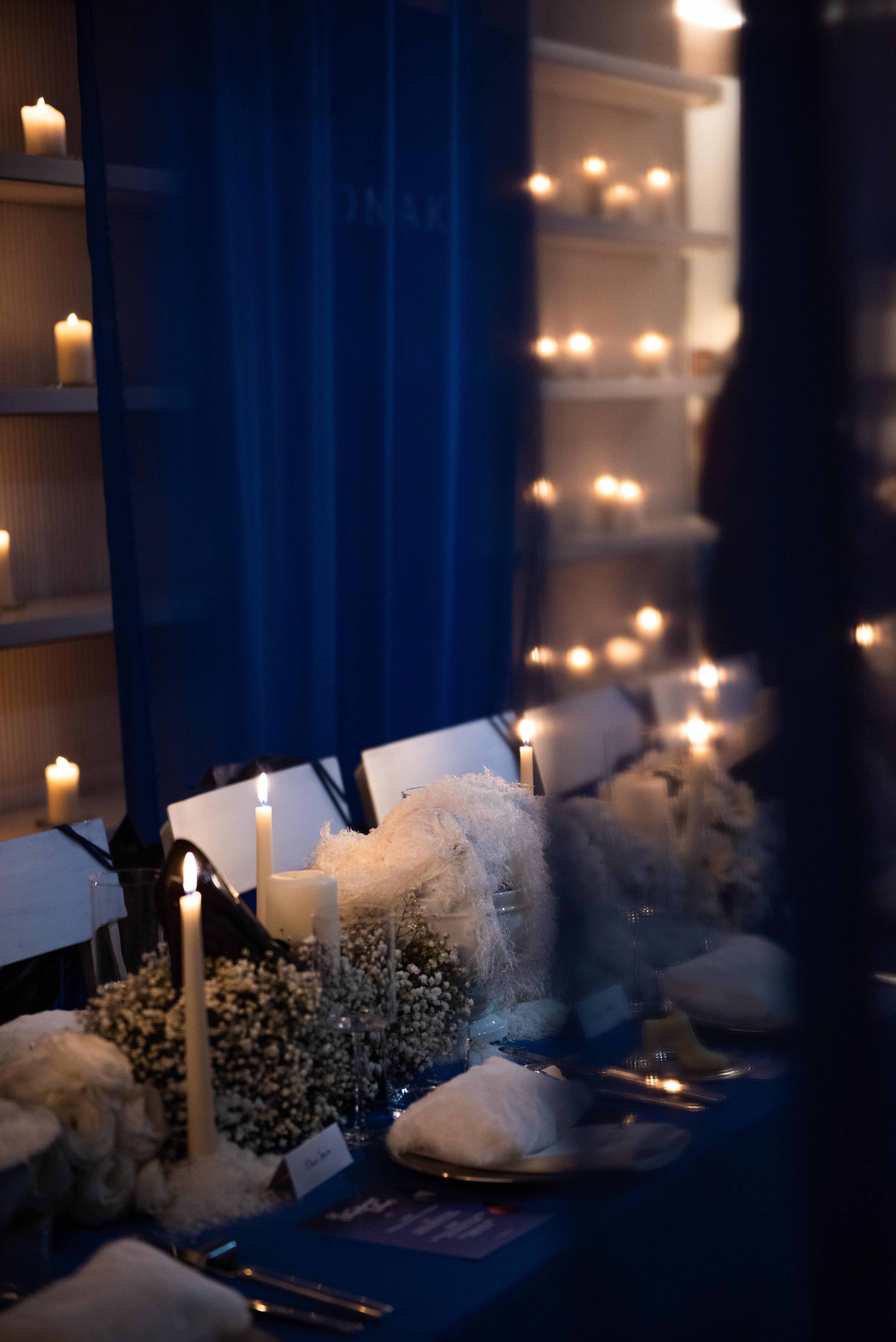Dinner table decorated with white flowers, candles, and white napkins in a dimly lit room with blue curtains and lit candles on shelves.