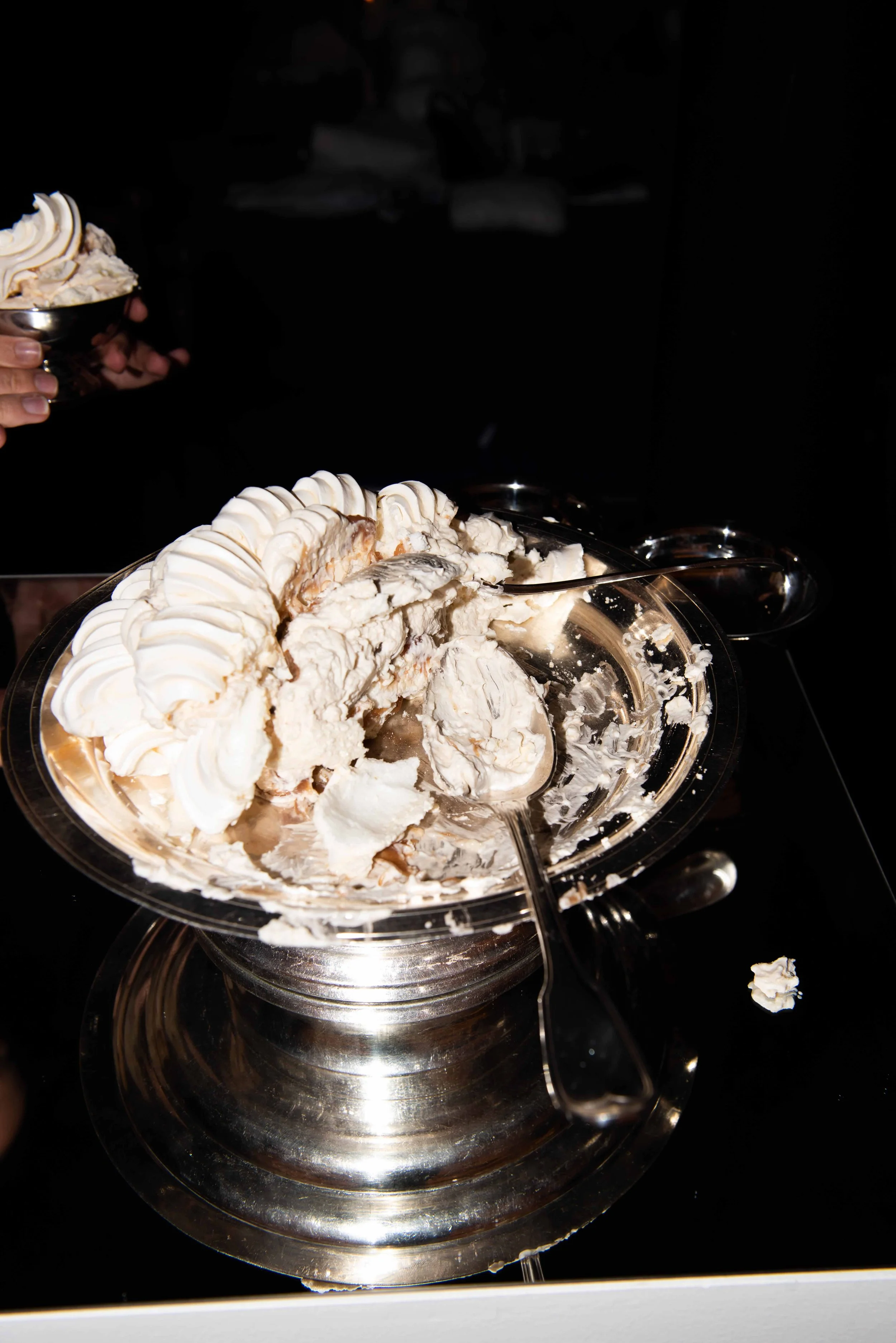 Ice cream in a large glass bowl, some blurred hands holding a smaller cup of ice cream, dark background.