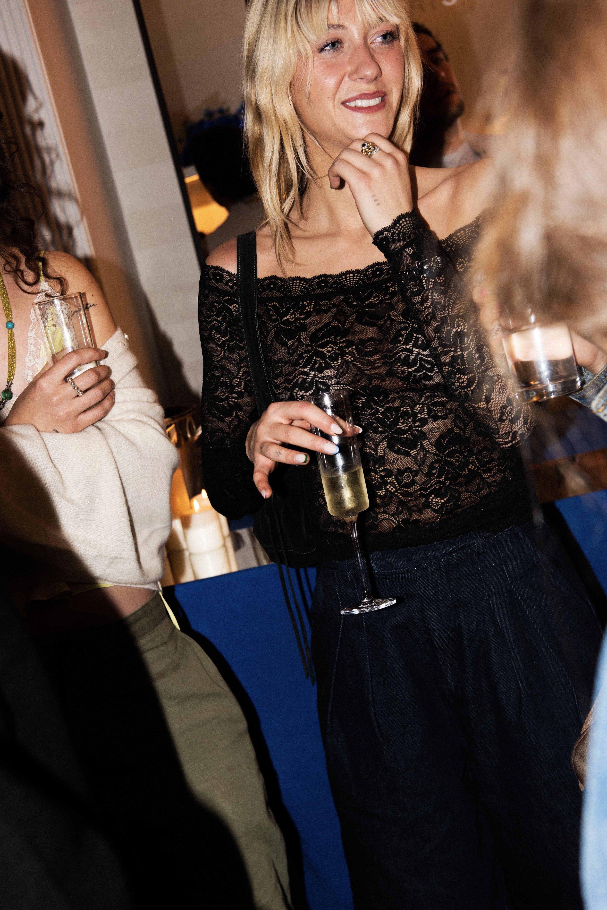 A woman with blonde hair in loose waves, wearing a black lace off-shoulder top and dark pants, holding a glass of champagne at a social gathering.