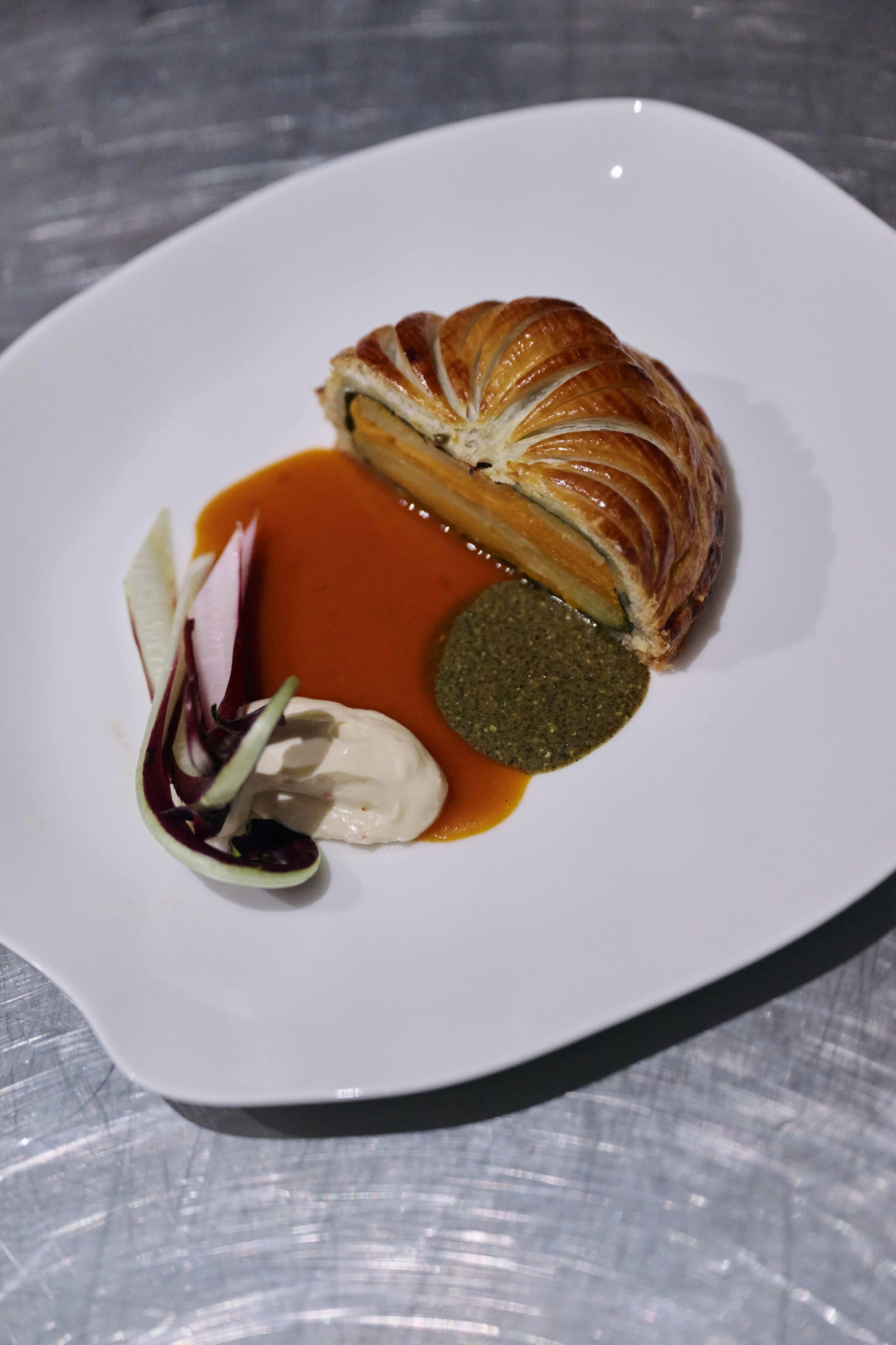 A plated pastry dish with a sliced baked pastry, green and black sauces, and a dollop of cream or sauce, garnished with thin slices of vegetables or fruit.