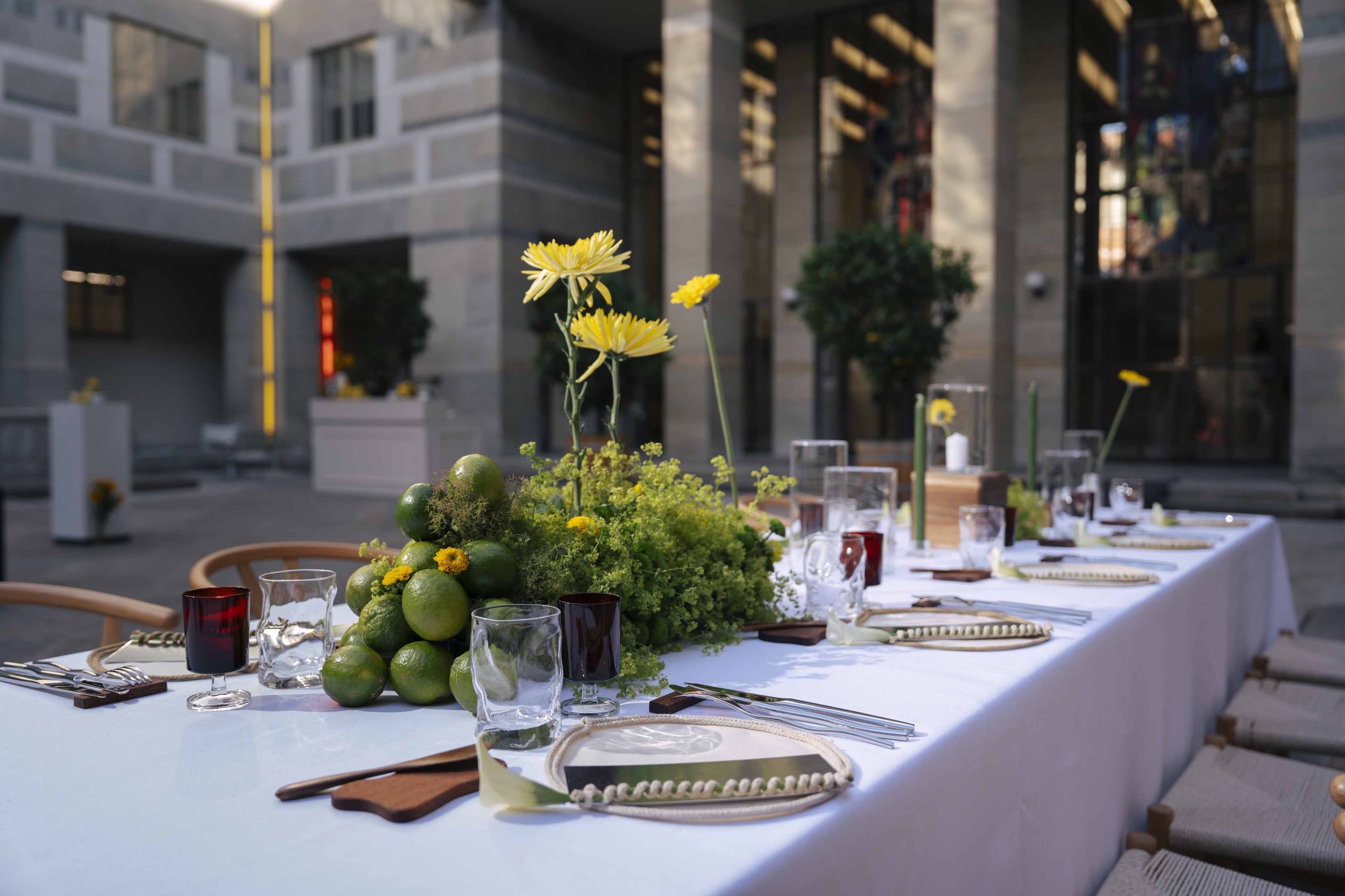 Long banquet table set outdoors with decorated centerpiece of green limes, yellow flowers, and greenery, surrounded by chairs and elegant glassware, in an urban courtyard with modern buildings in the background.