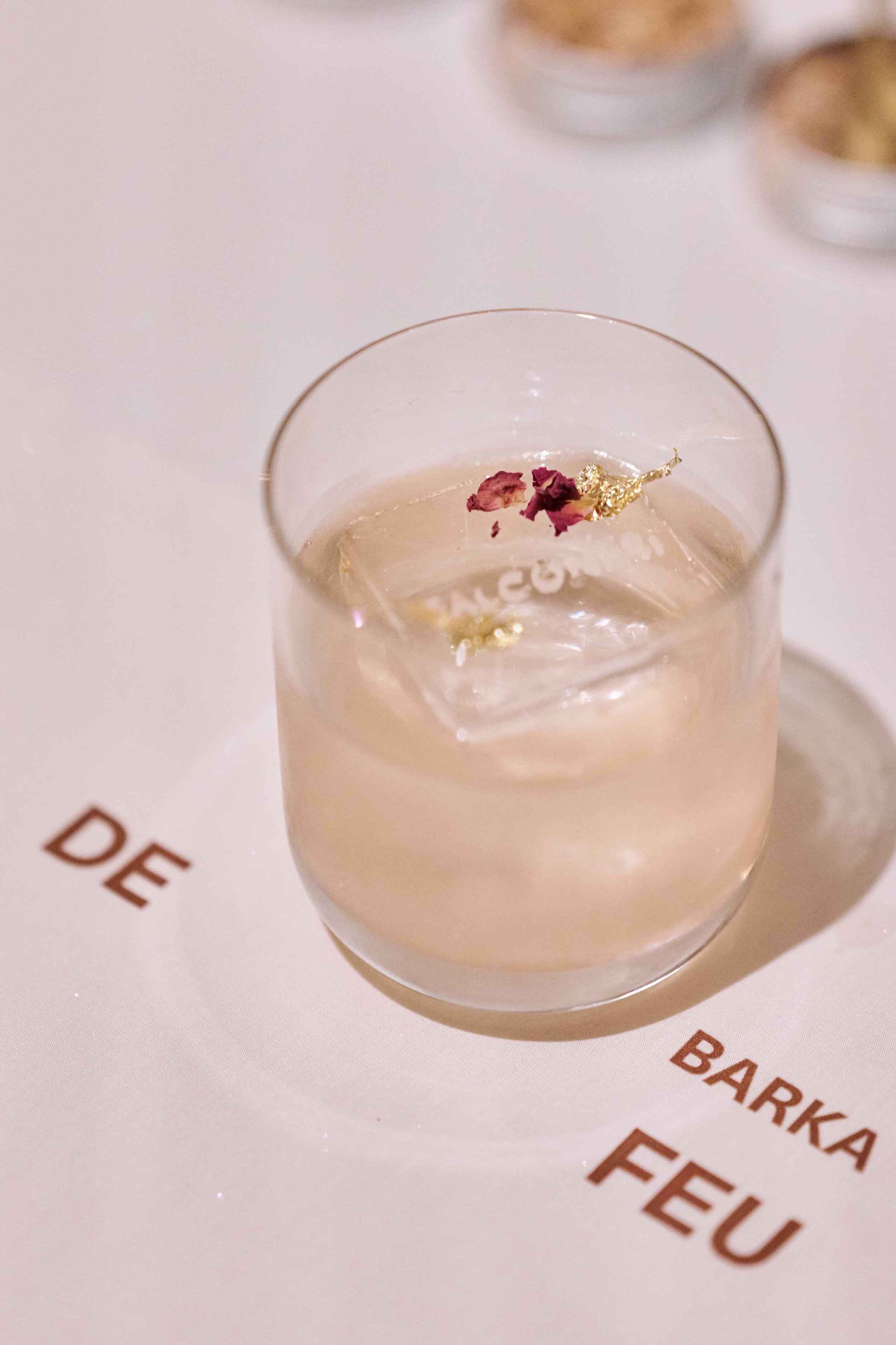 A glass of water with ice and edible flower petals, placed on a menu or table surface with the words 'DE', 'BARKA', 'FEU' visible.