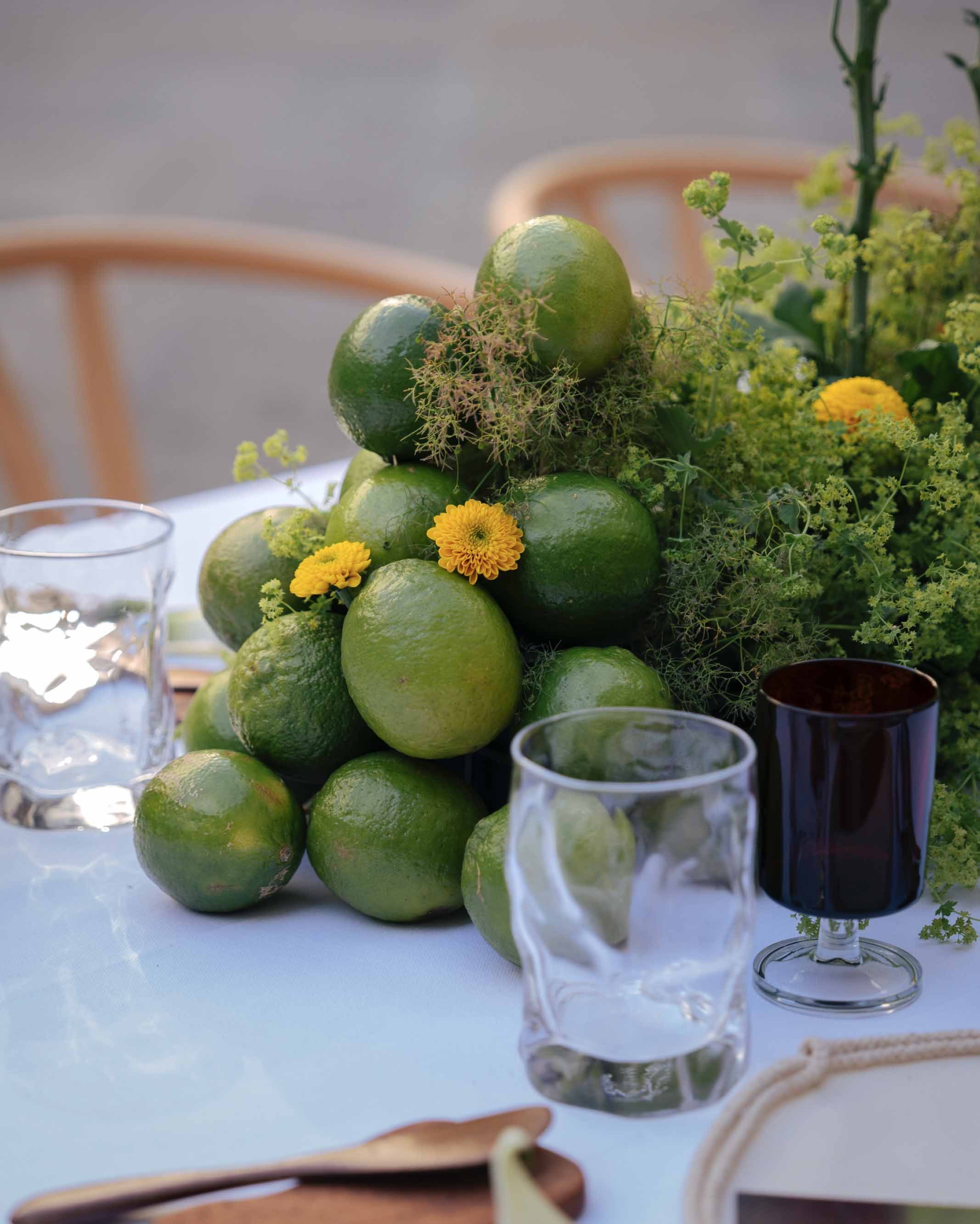Lime centerpiece with yellow flowers and greenery on a white table, with glassware and wooden utensils nearby.