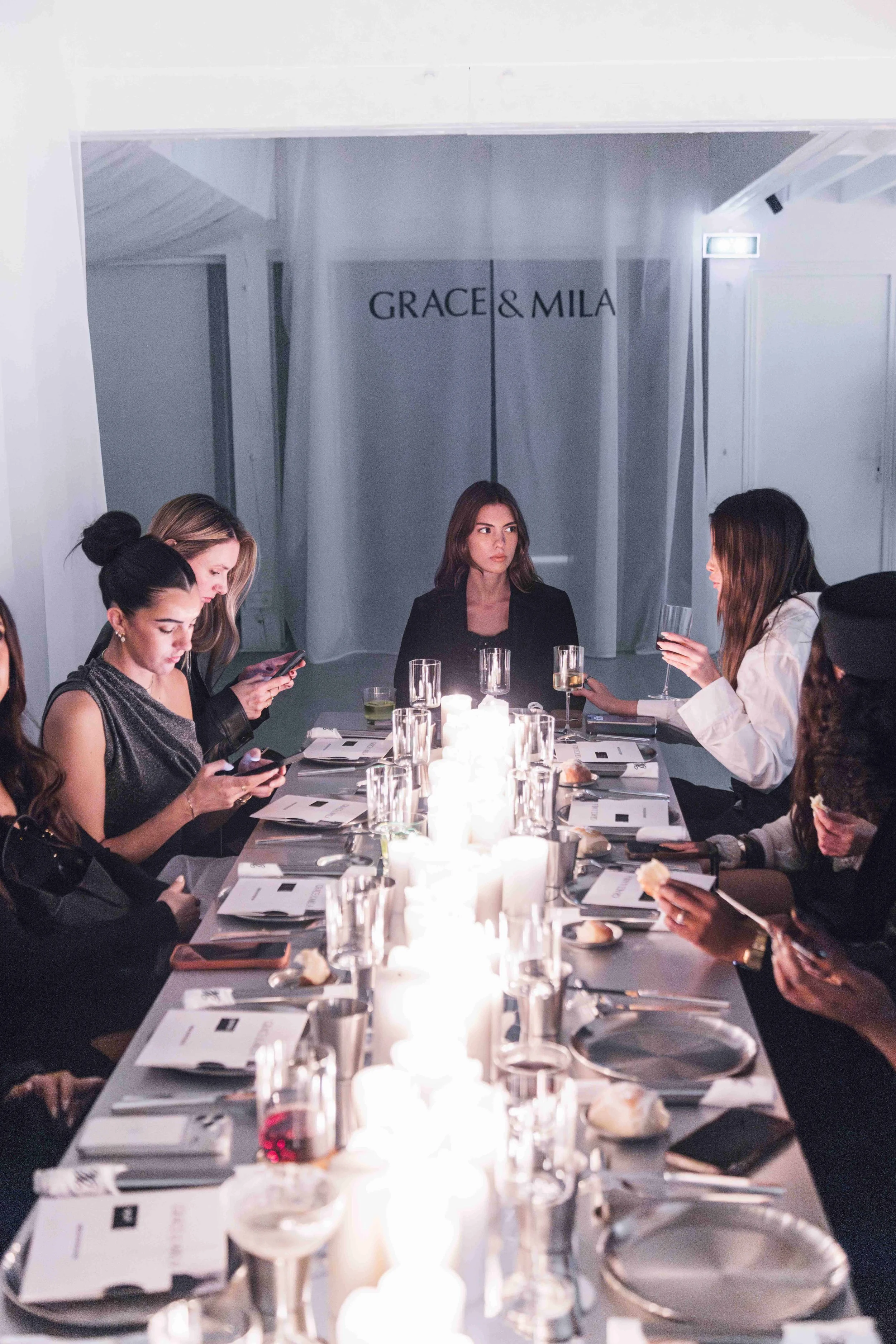 Women sitting around a long dining table with candles, drinking glasses, and plates, with a backdrop that says 'GRACE & MILA'.