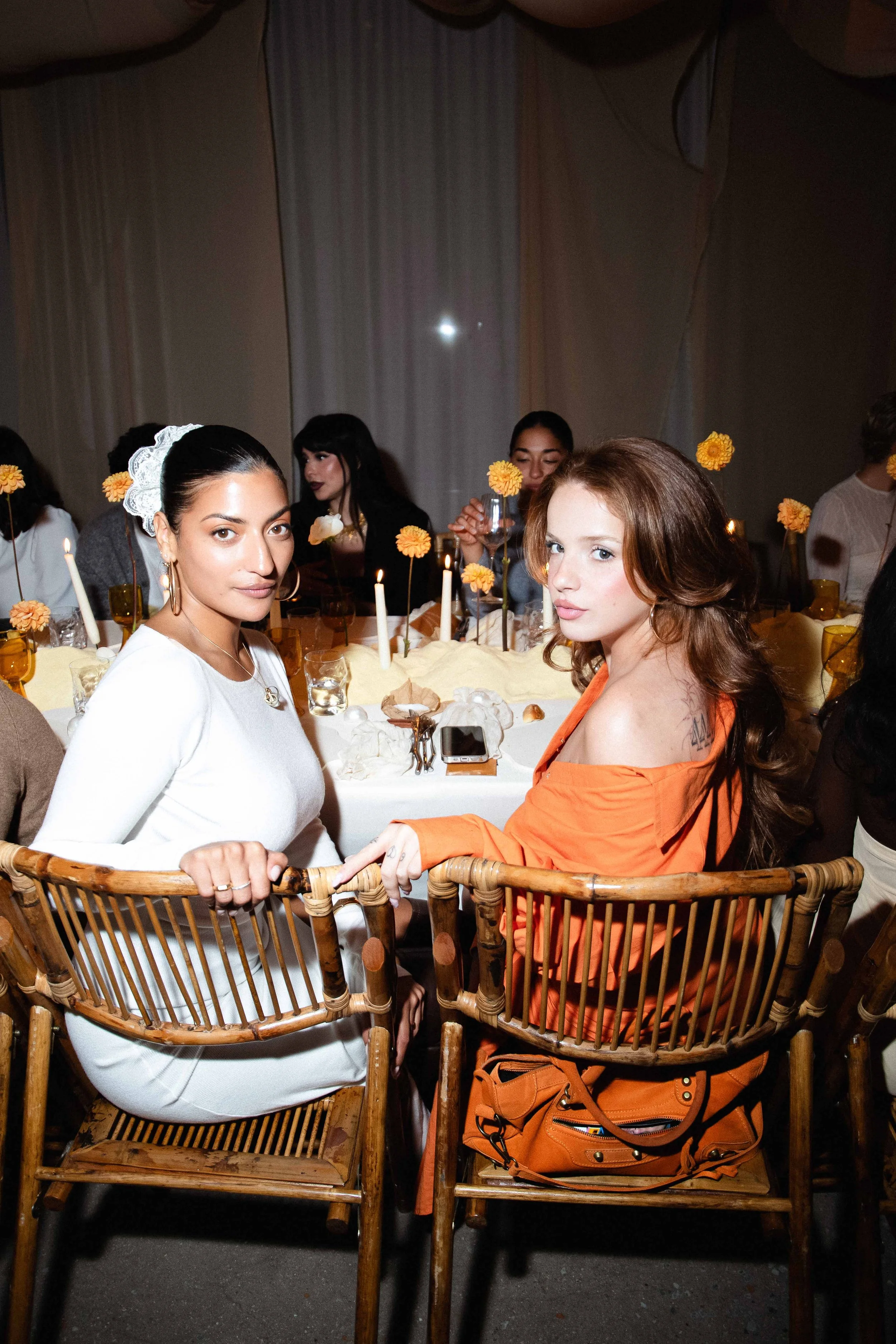 Two women sitting at a dining table during a gathering or celebration, with other people in the background, decorated with candles and orange flowers.