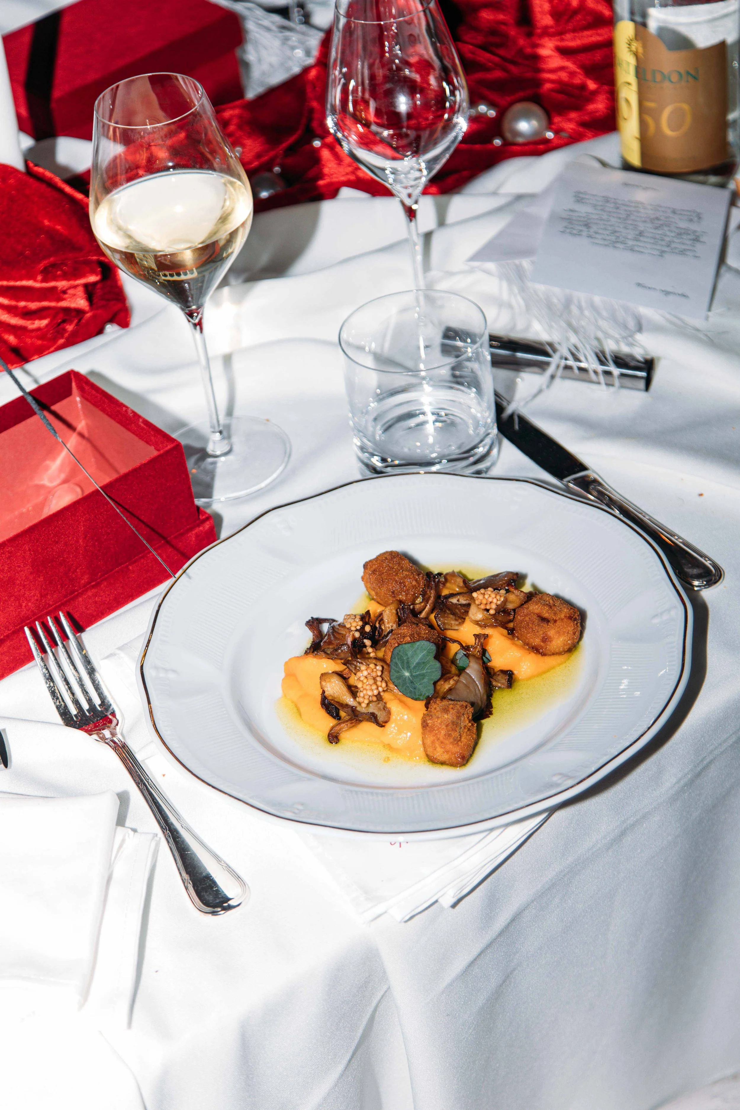 A fine dining table setting with a white plate of gourmet food, wine glasses, a red gift box, and a bottle of champagne on a white tablecloth.