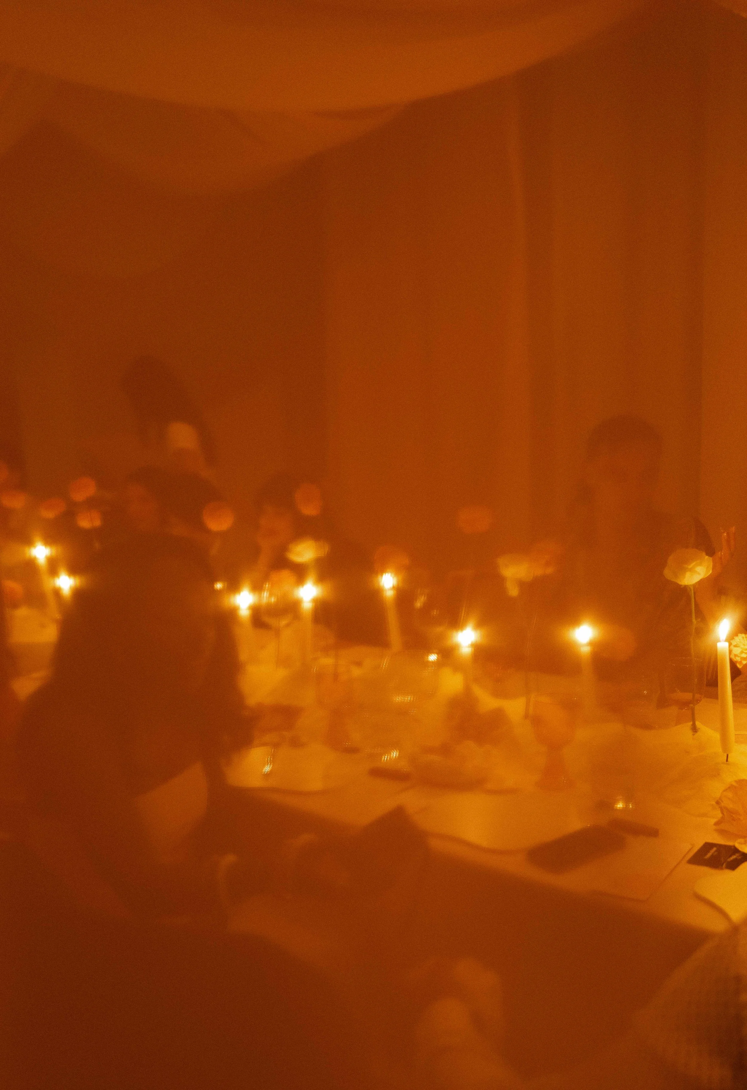 A dimly lit indoor scene with a table set with food, glasses, and lit candles, where people are gathered and appear to be praying or observing a religious ritual.