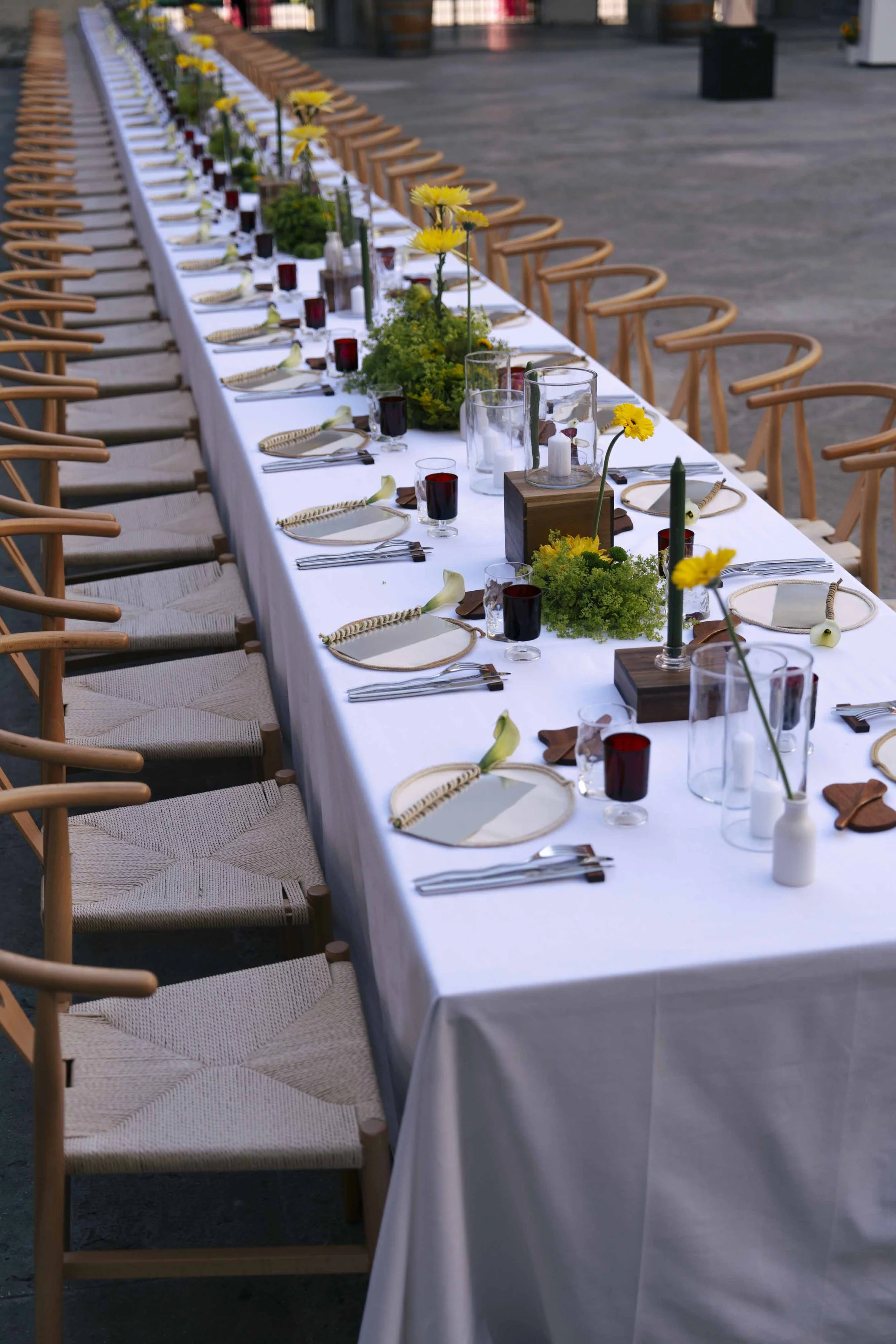 Long banquet table decorated with white tablecloth, yellow flowers, and candles, set for an outdoor event with wooden chairs evenly spaced around it.