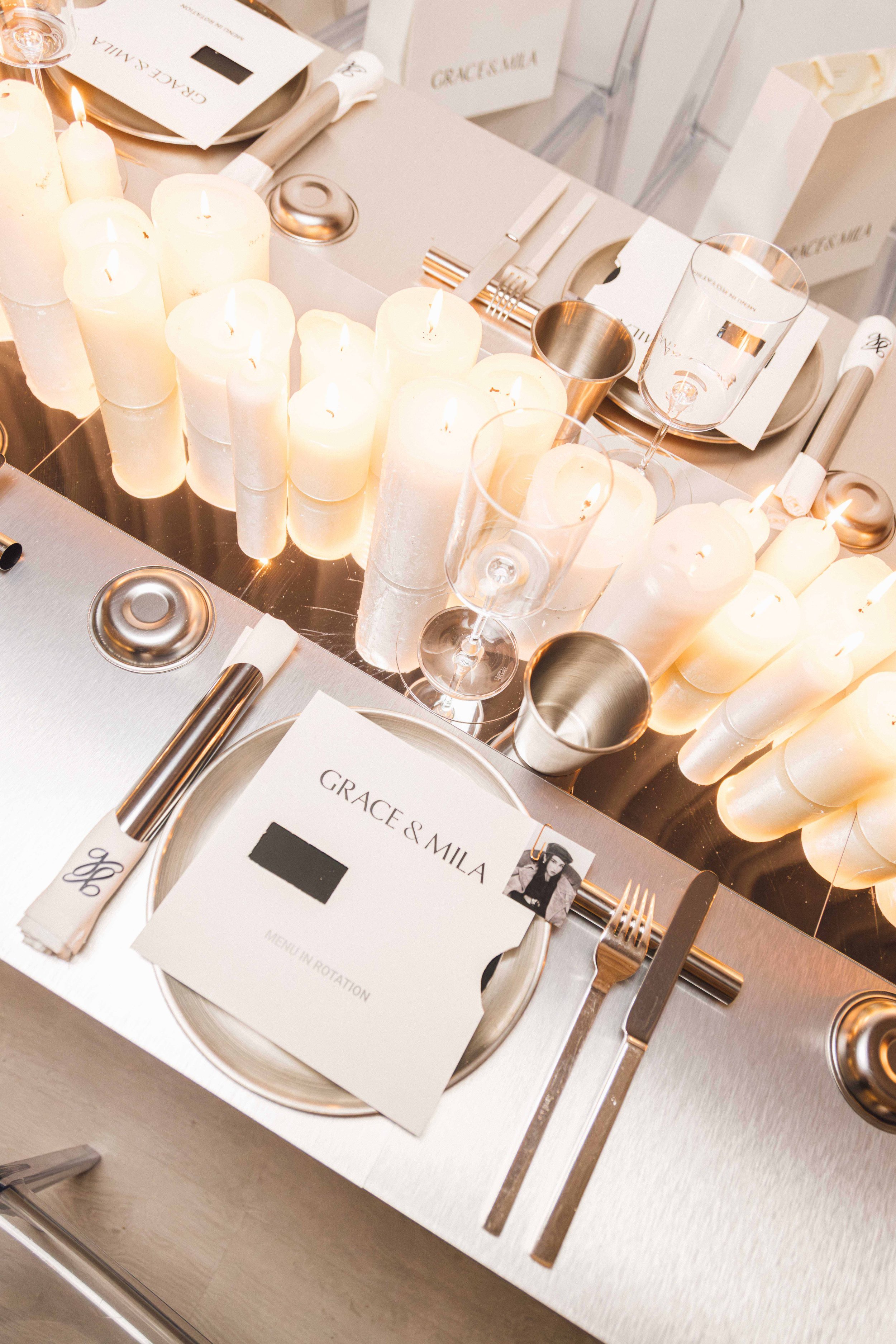 Elegant event table setup featuring a centerpiece of white candles, with place settings including a menu card labeled 'Grace & Mila', utensils, a glass of water, and a wine glass, on a silver table.
