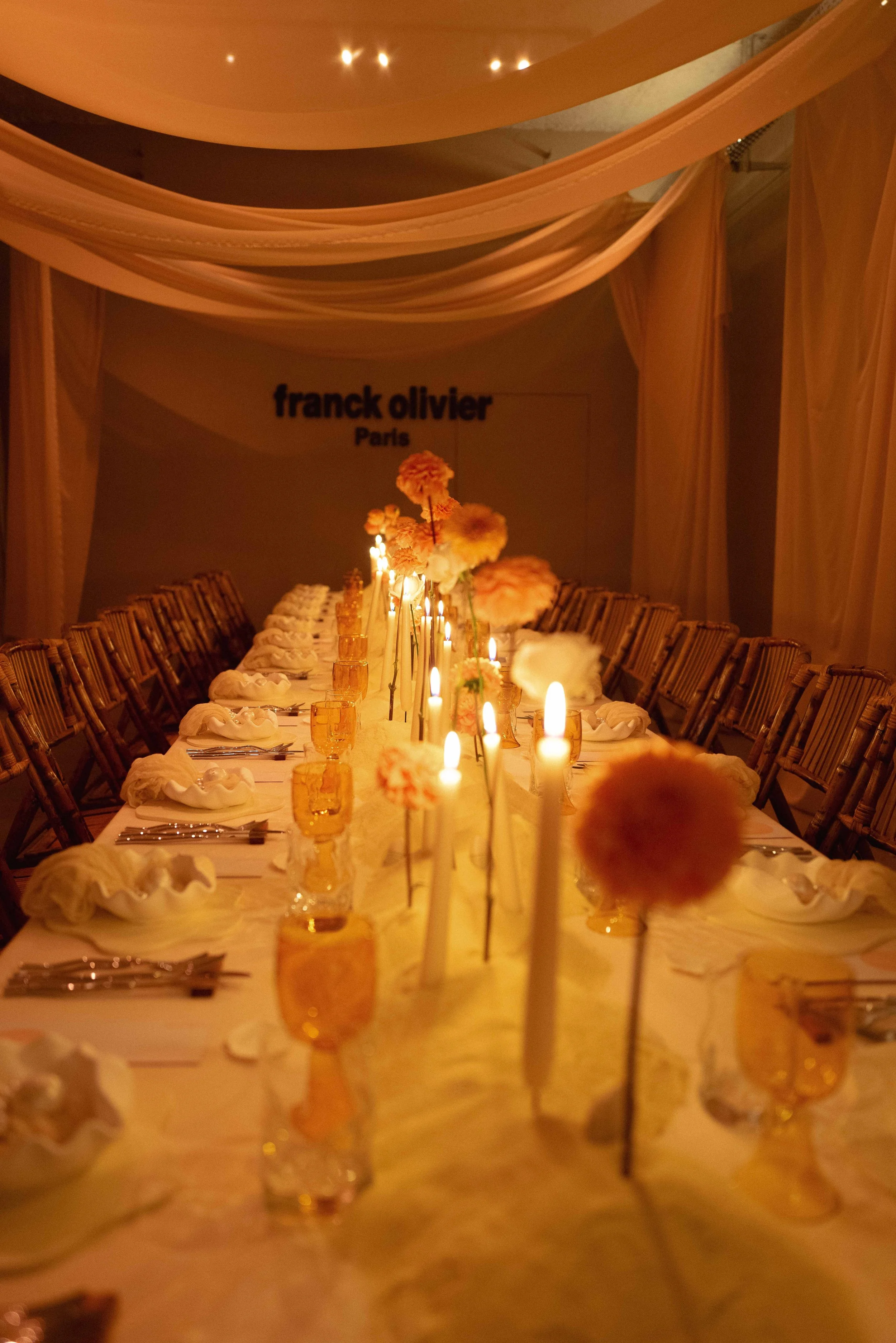 Elegant dinner table set with candles and floral centerpieces in a softly lit room with draped fabric and a 'franck olivier Paris' sign on the wall.