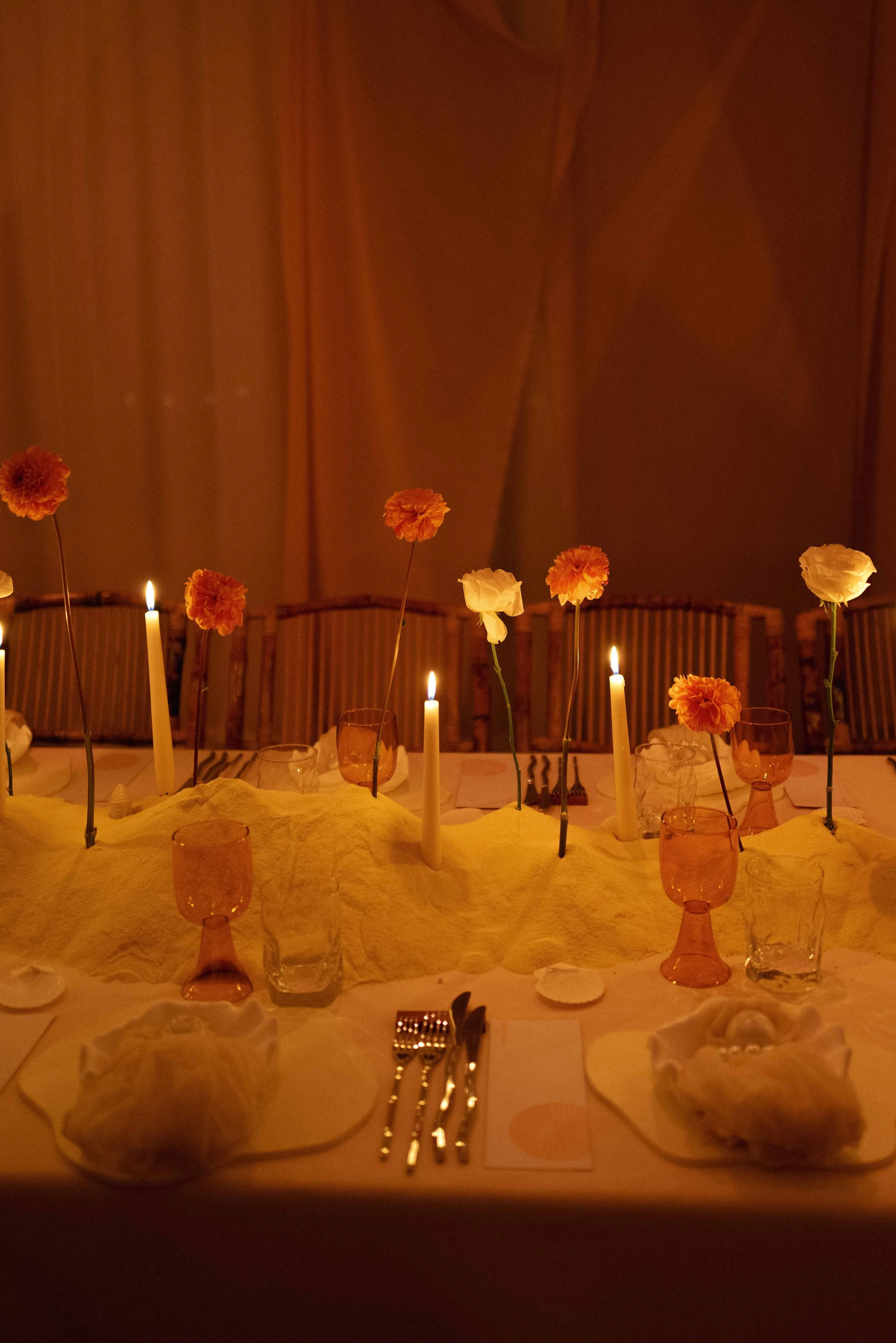 A dimly lit table decorated with pink and white flowers, candles, glasses, and tableware, set for a formal dinner.