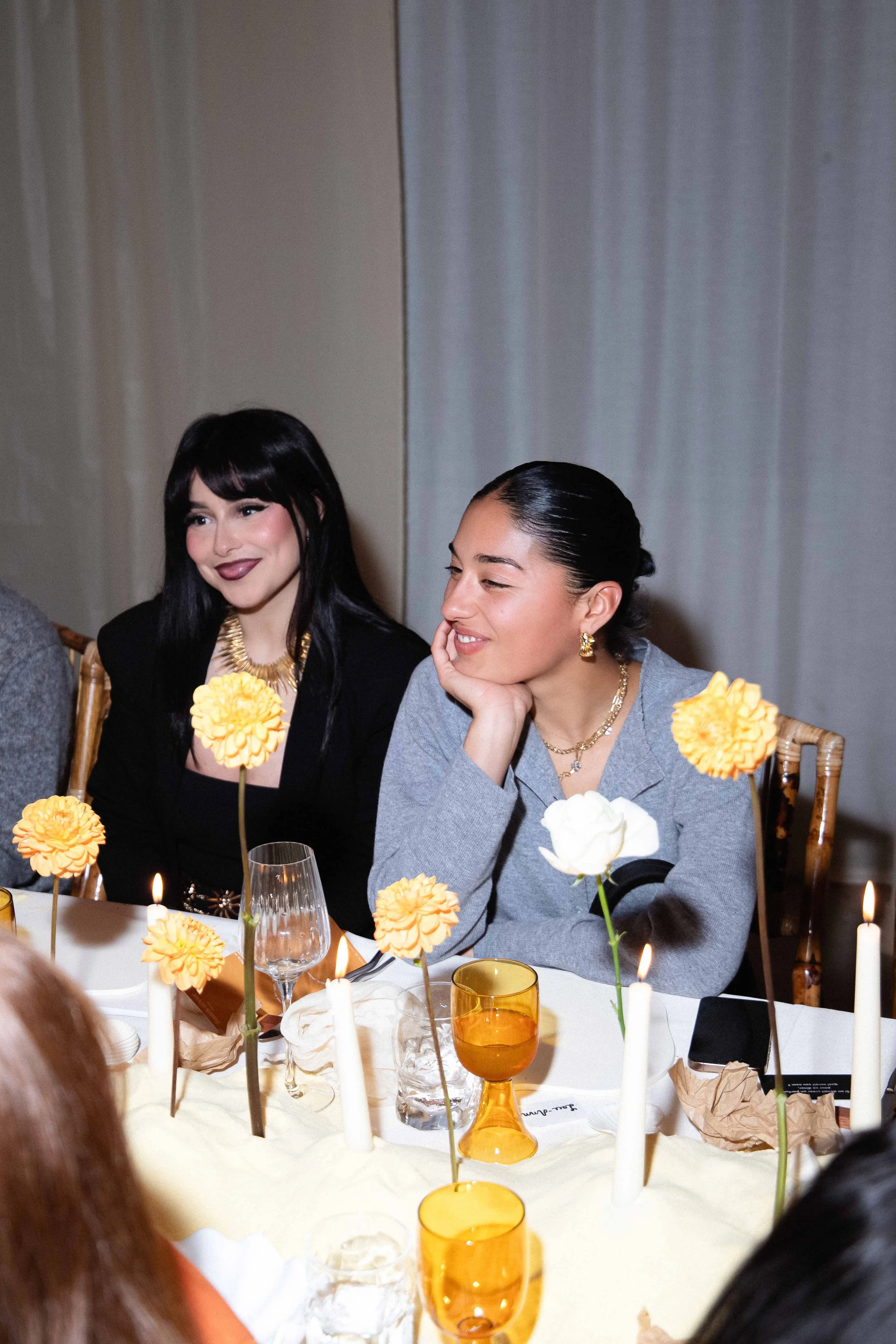 Two women sitting at a decorated table with candles and flowers, smiling during a celebration.
