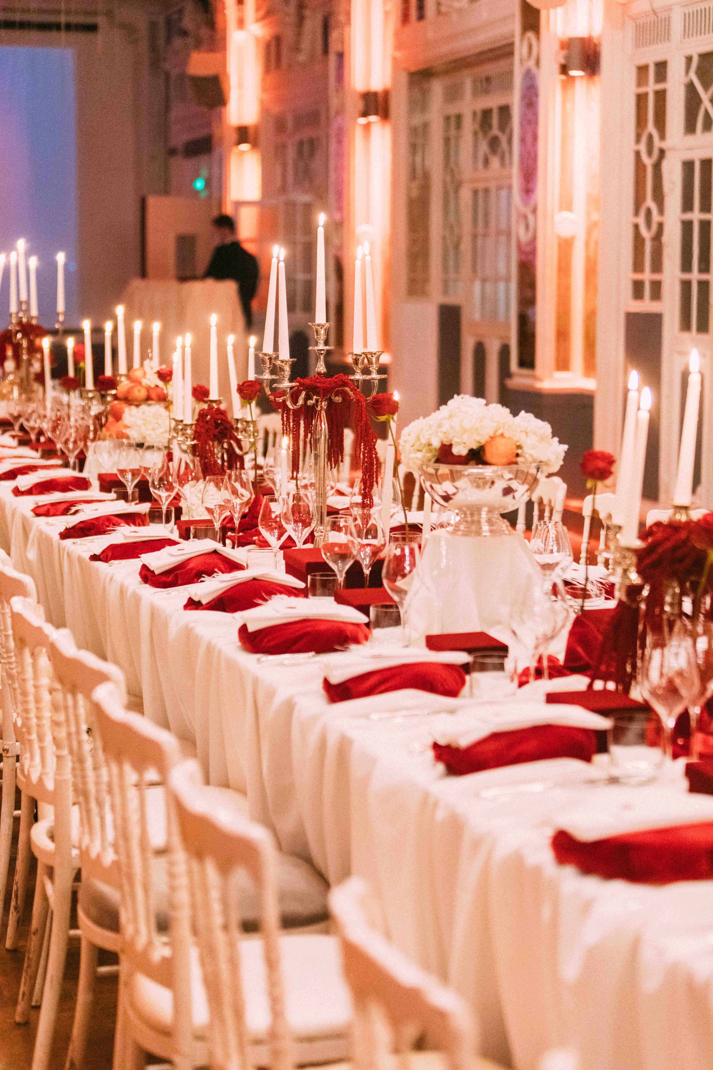 Elegant banquet table decorated with white tablecloths, red napkins, floral centerpieces, and tall candle holders with lit white candles, set in a beautifully lit room for a formal event.