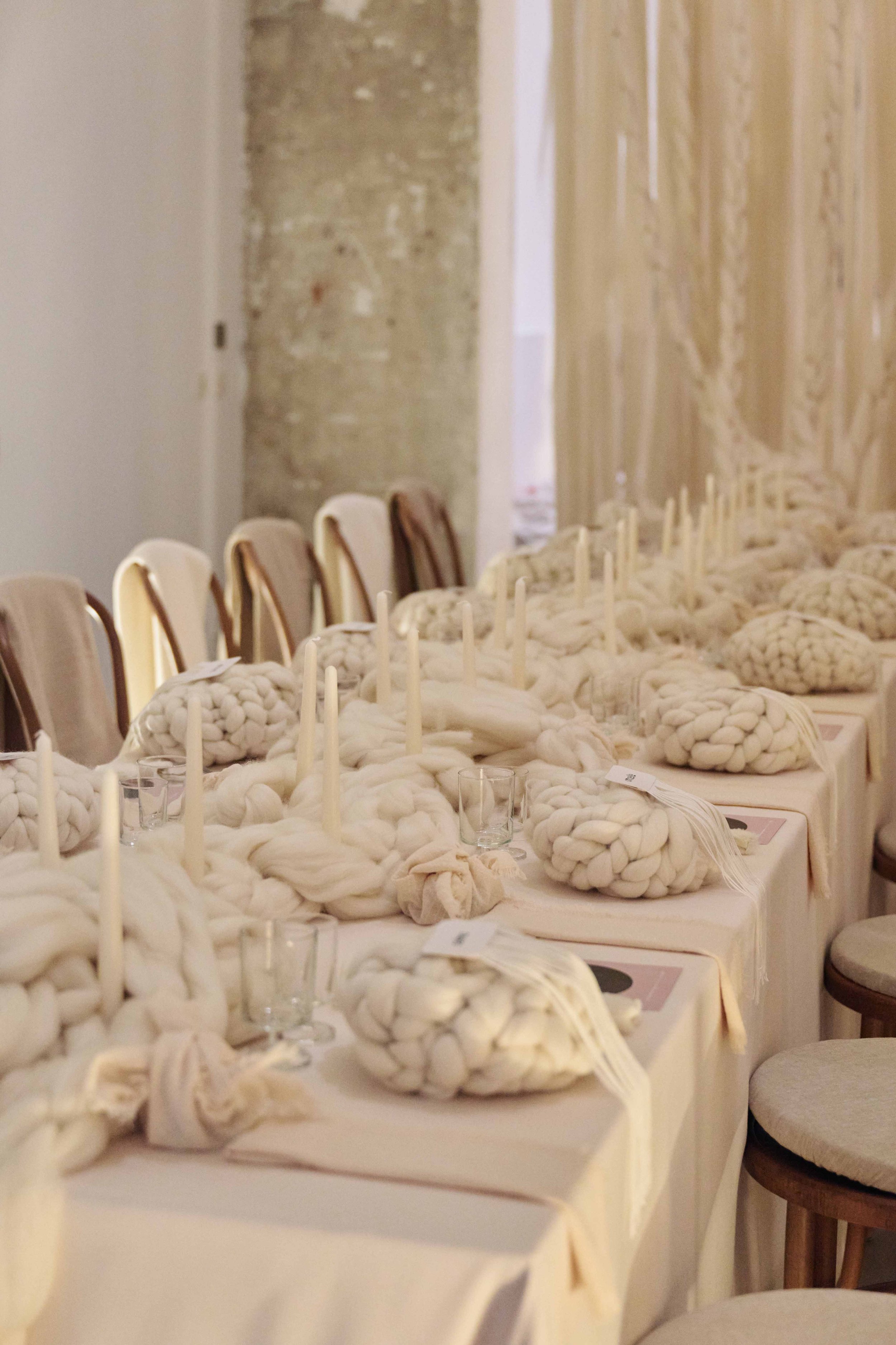 A long dining table set with white tablecloths, featuring large knotted wool centerpieces, candles, and glassware, in a rustic and minimalist style.