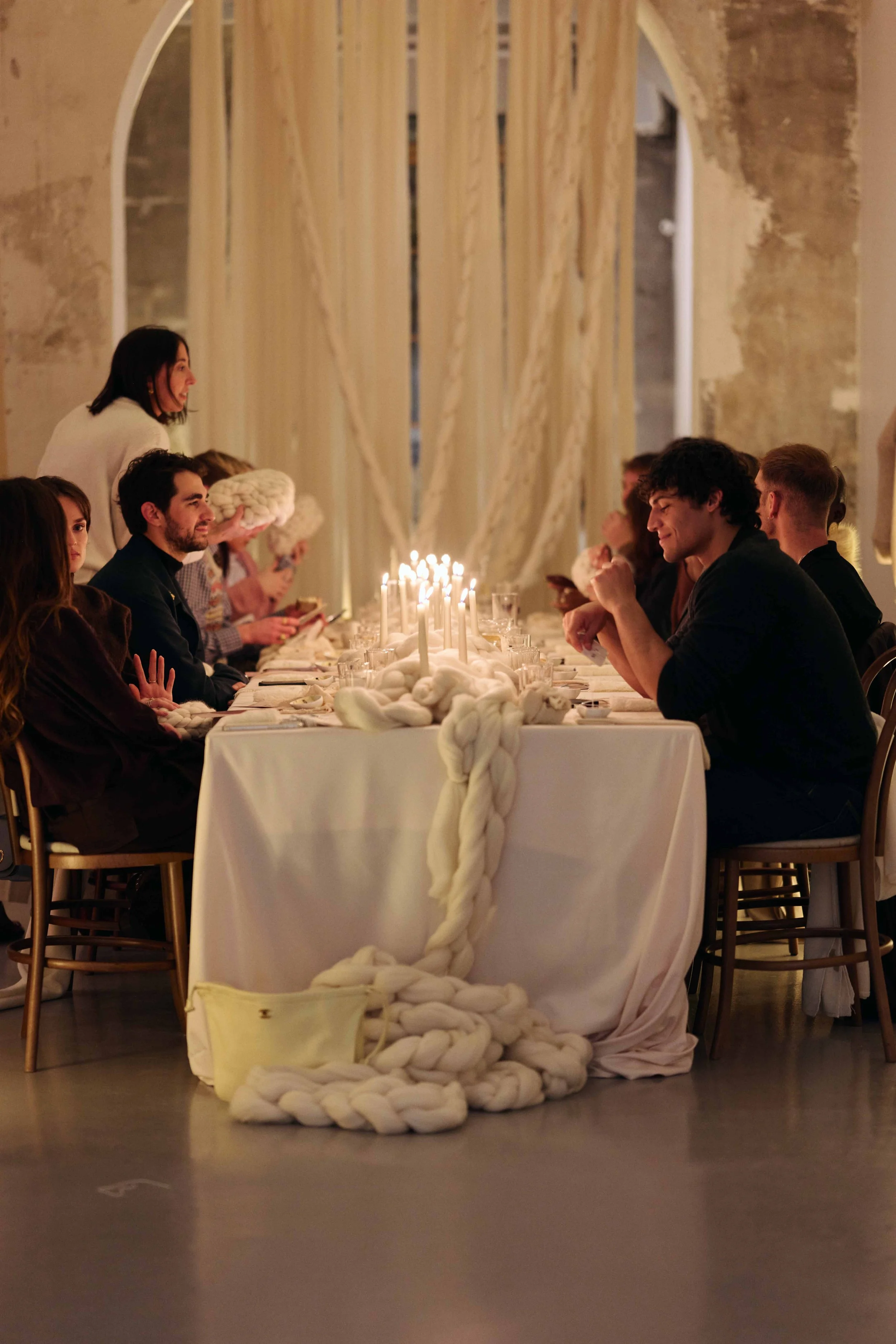 People sitting at a decorated dinner table with a white cloth, white candles, and a large braided white cloth centerpiece, during an indoor event with dim lighting.