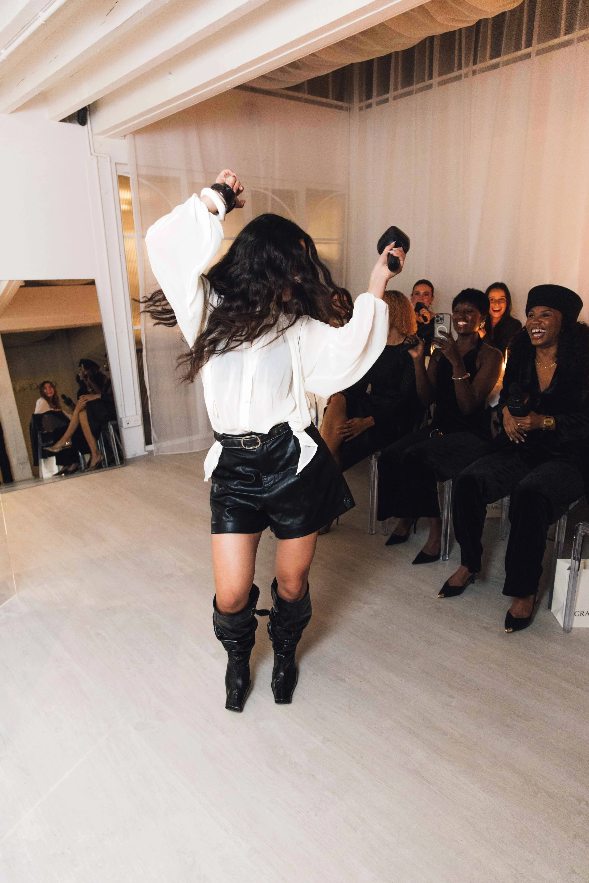 A woman with long dark wavy hair dancing or singing with her arms raised, wearing a white blouse and black leather shorts with high gray boots, in front of a group of seated women who are smiling and holding microphones and phones.