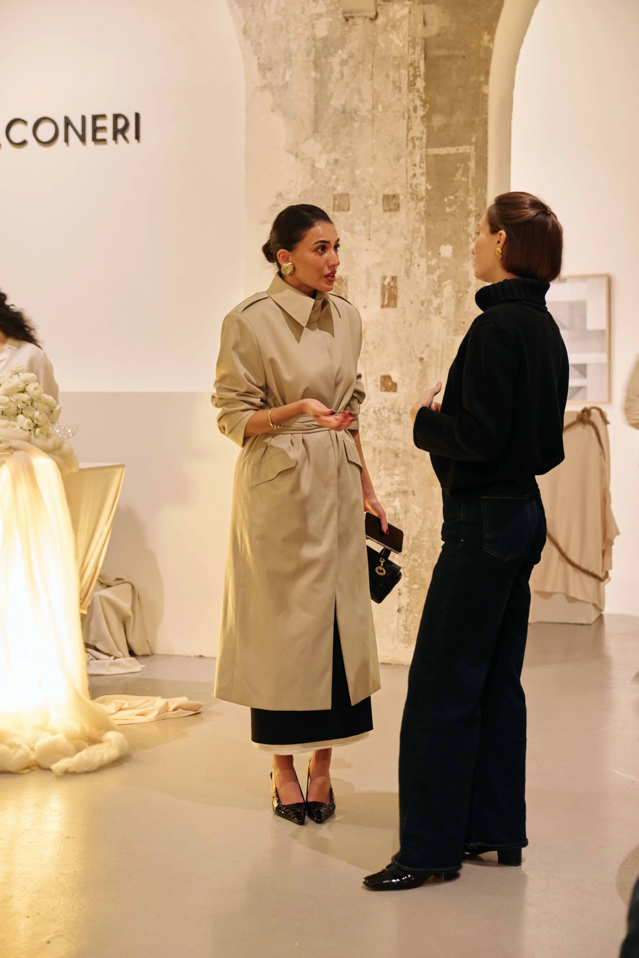 Two women engaged in conversation at an art gallery or exhibit. One wears a beige trench coat and high heels, the other is dressed in a black turtleneck and dark jeans.
