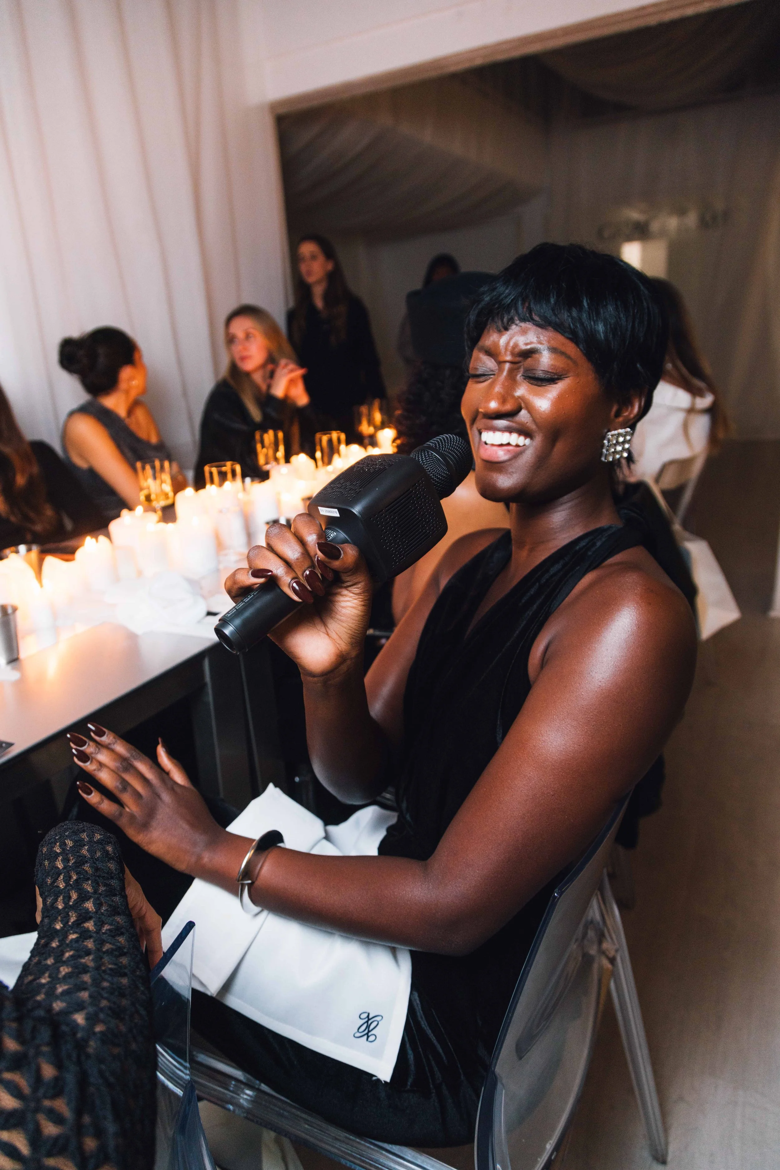 A woman with short black hair singing into a microphone during a dinner event, with a table of lit candles and other guests in the background.