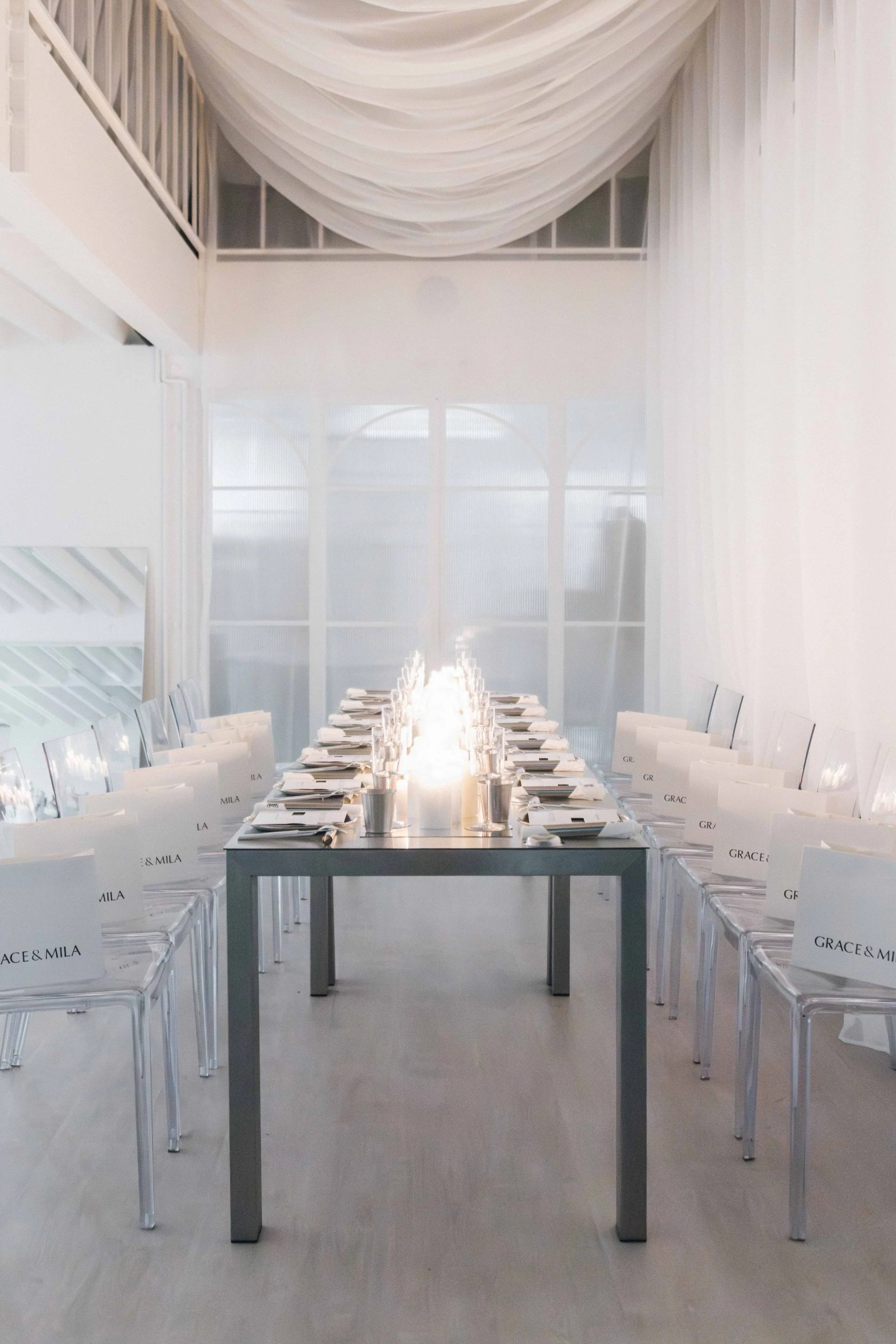 Elegant, minimalist event space with a long table set for a formal gathering, surrounded by clear acrylic chairs with name cards, and a soft lighting installation on the table, with white curtains and large windows creating a bright, airy atmosphere.