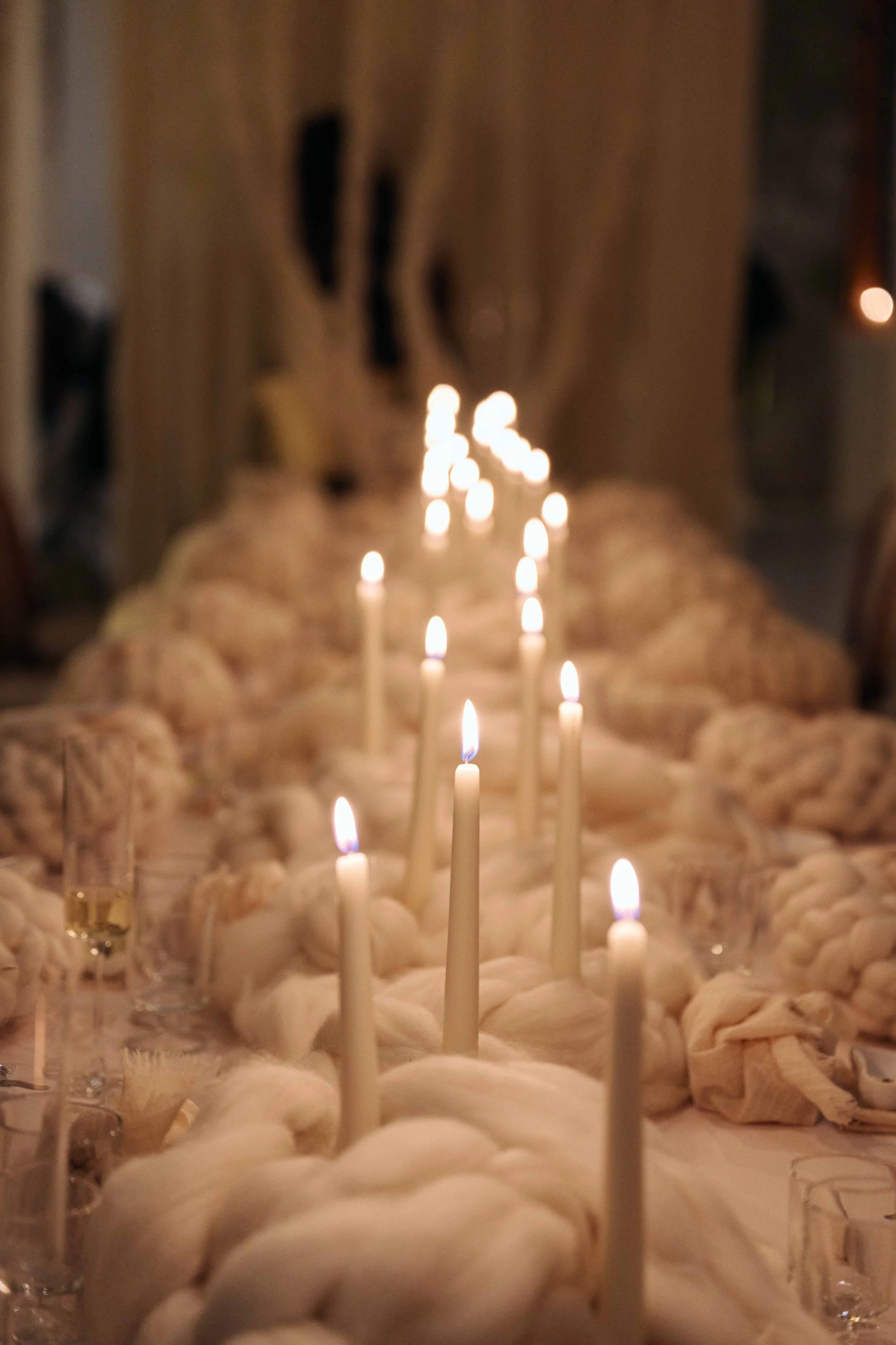 A dining table decorated with tall white candles lit on braided white fabric, with wine glasses and a champagne flute, and warm ambient lighting.