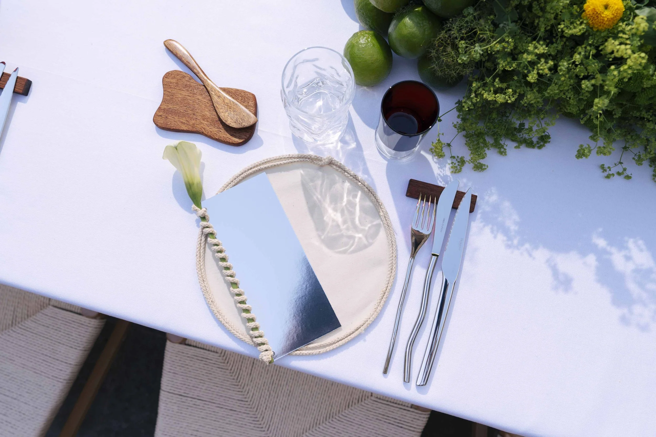 A table setting with a mirror plate on a beige cloth napkin with a floral decoration, stainless steel fork, knife, and spoon, a glass of water, a glass of red wine, a small wooden cutting board with a wooden spoon, and a bunch of green limes and greenery on a white tablecloth.