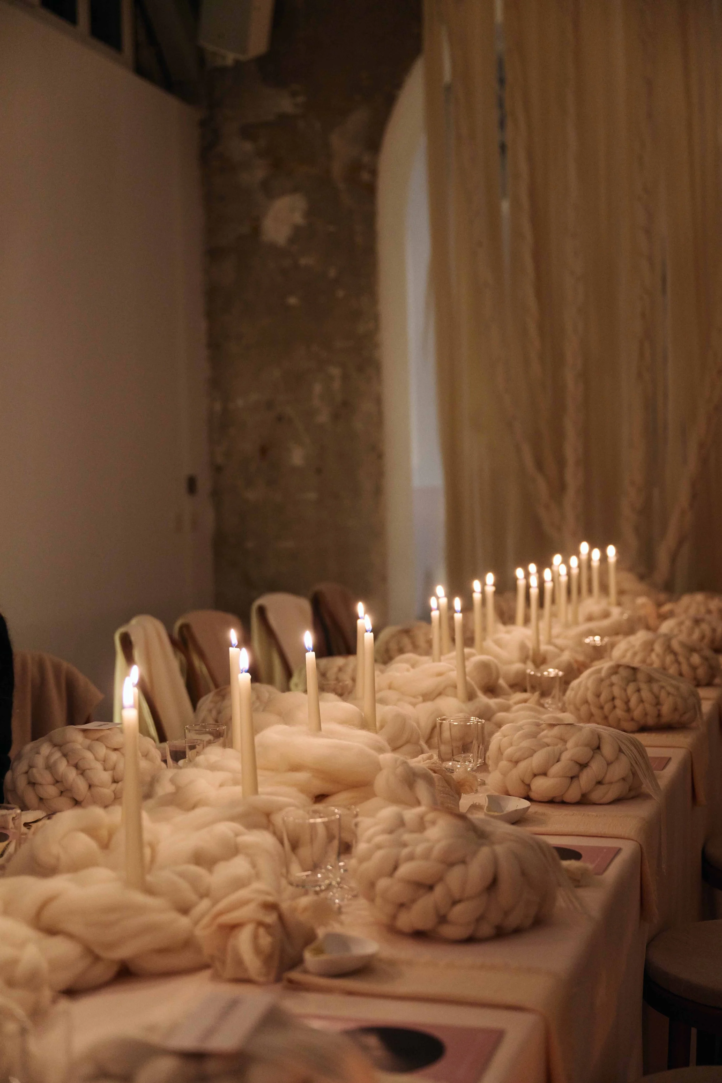 Long dining table decorated with large knitted cream-colored tablecloths, white candles lit, in an industrial-style room with exposed concrete wall