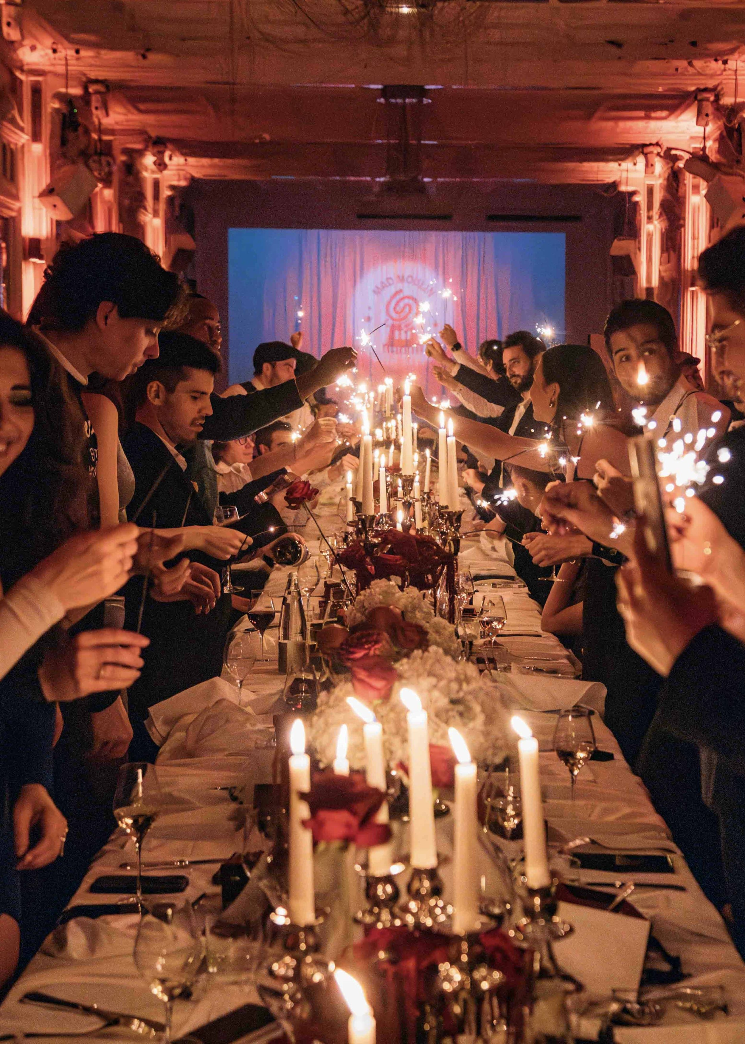 A large dinner party with guests holding sparklers in a dimly lit room, decorated with candles and floral arrangements, celebrating around a long table.
