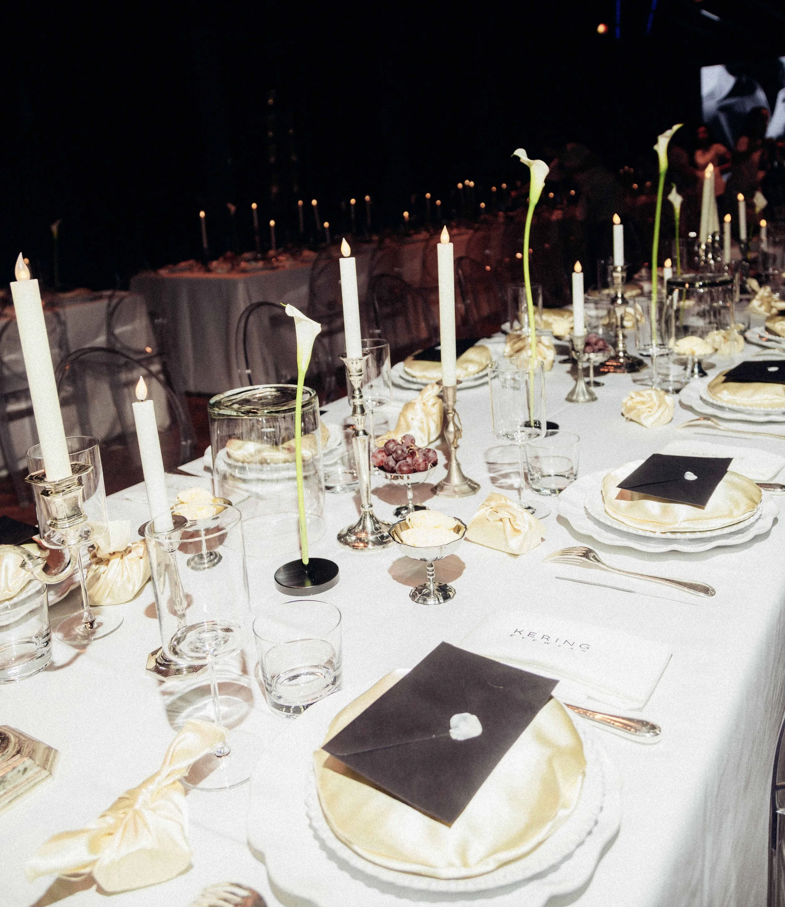 Evening banquet table set with white tablecloth, candles, calla lilies, glasses, and black and white menus.