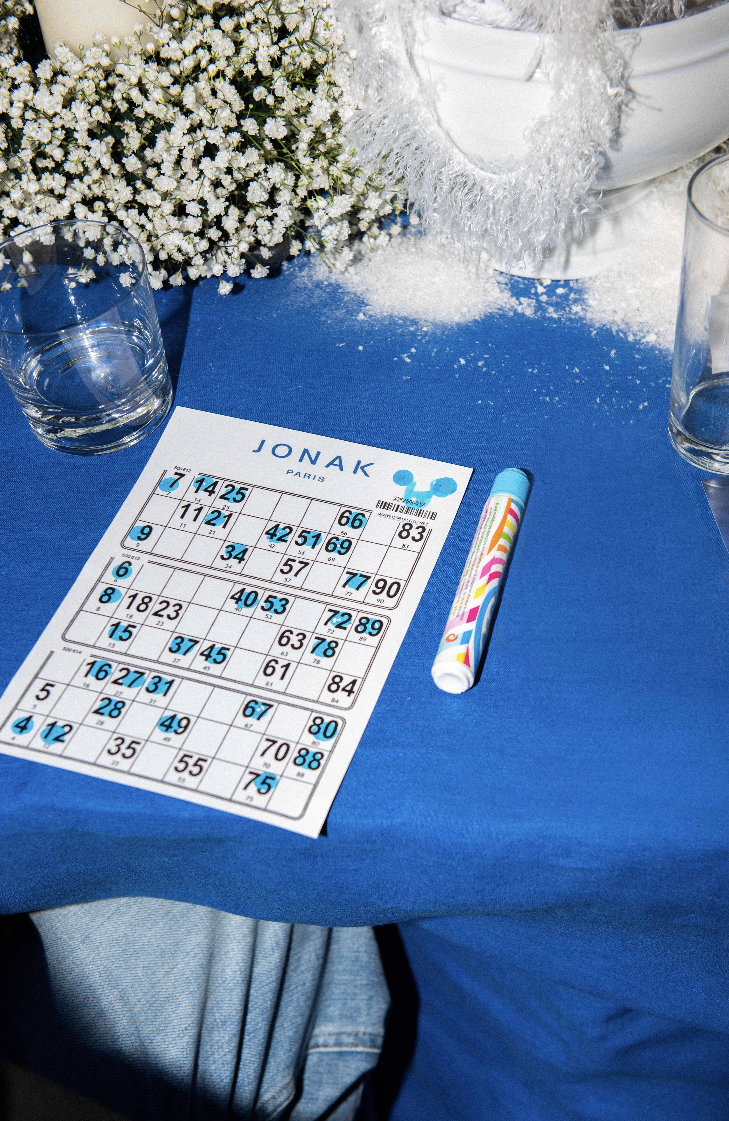 A bingo card titled 'JONAK PARIS' on a blue tablecloth, with a colorful marker, clear glass of water, white flowers, and a decorative white bucket in the background.