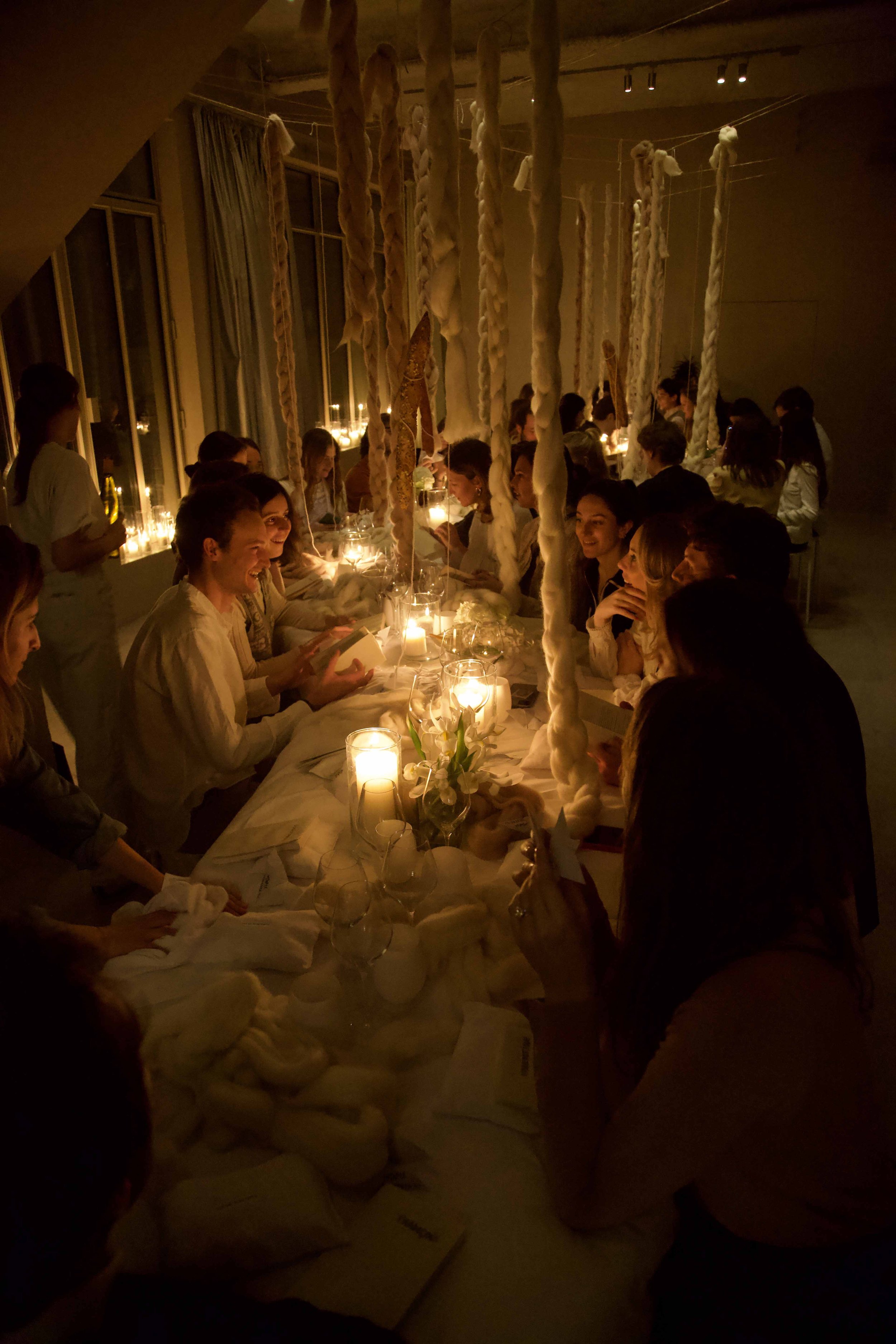 A dimly lit indoor dinner event with a long table decorated with candles, flowers, and cloth napkins. People are seated on both sides, engaging in conversation.