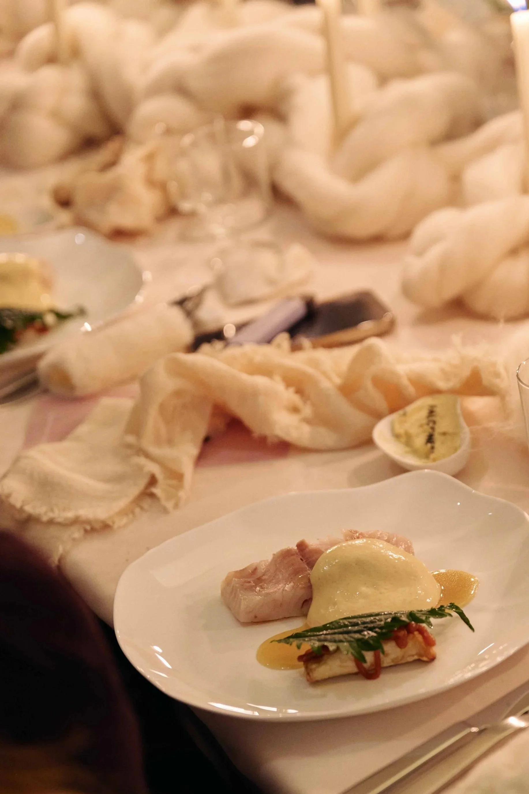 A plate with pieces of chicken, cheese, and herbs on a dinner table, with various bread rolls and a glass in the background.