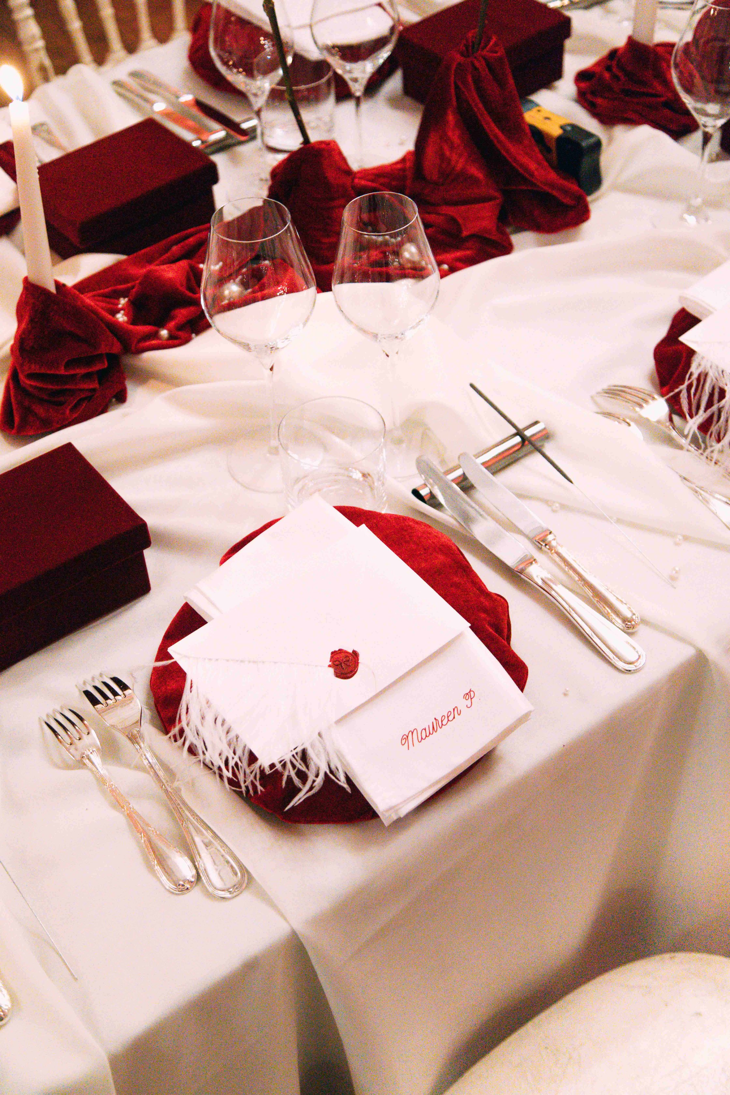 A formal dining table decorated for an event with white tablecloths, red napkins, and red gift boxes. Place settings include silverware, wine glasses, and a white envelope with a red wax seal and a name card labeled 'Maureen D.' on a red napkin.