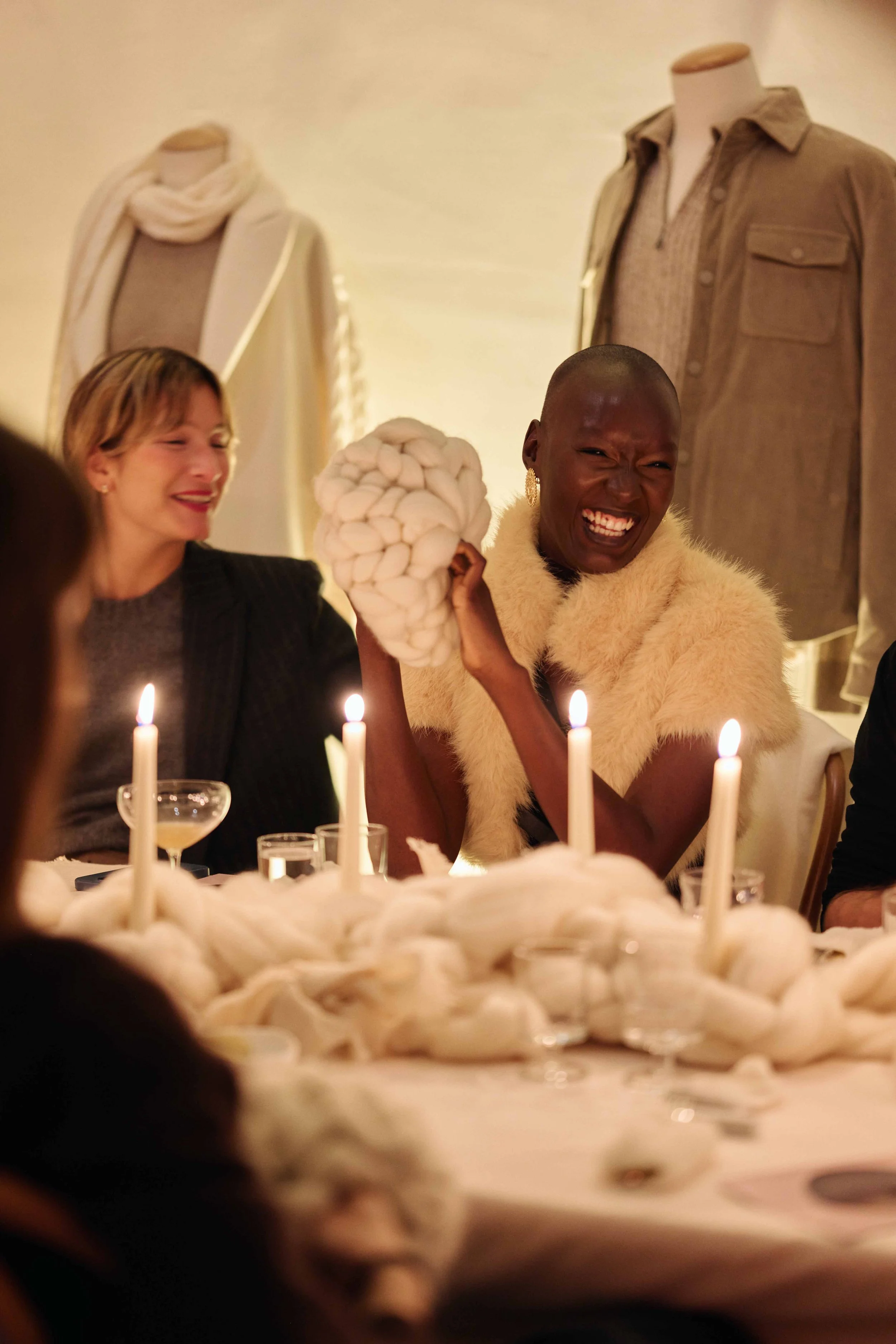 A woman with a shaved head wearing a cream-colored fur stole is smiling and holding a large bundle of white yarn or wool at a dinner party. She is seated at a table with lit candles and other guests, with oddly styled white decorations on the table. In the background, there are mannequins dressed in neutral-toned clothing.
