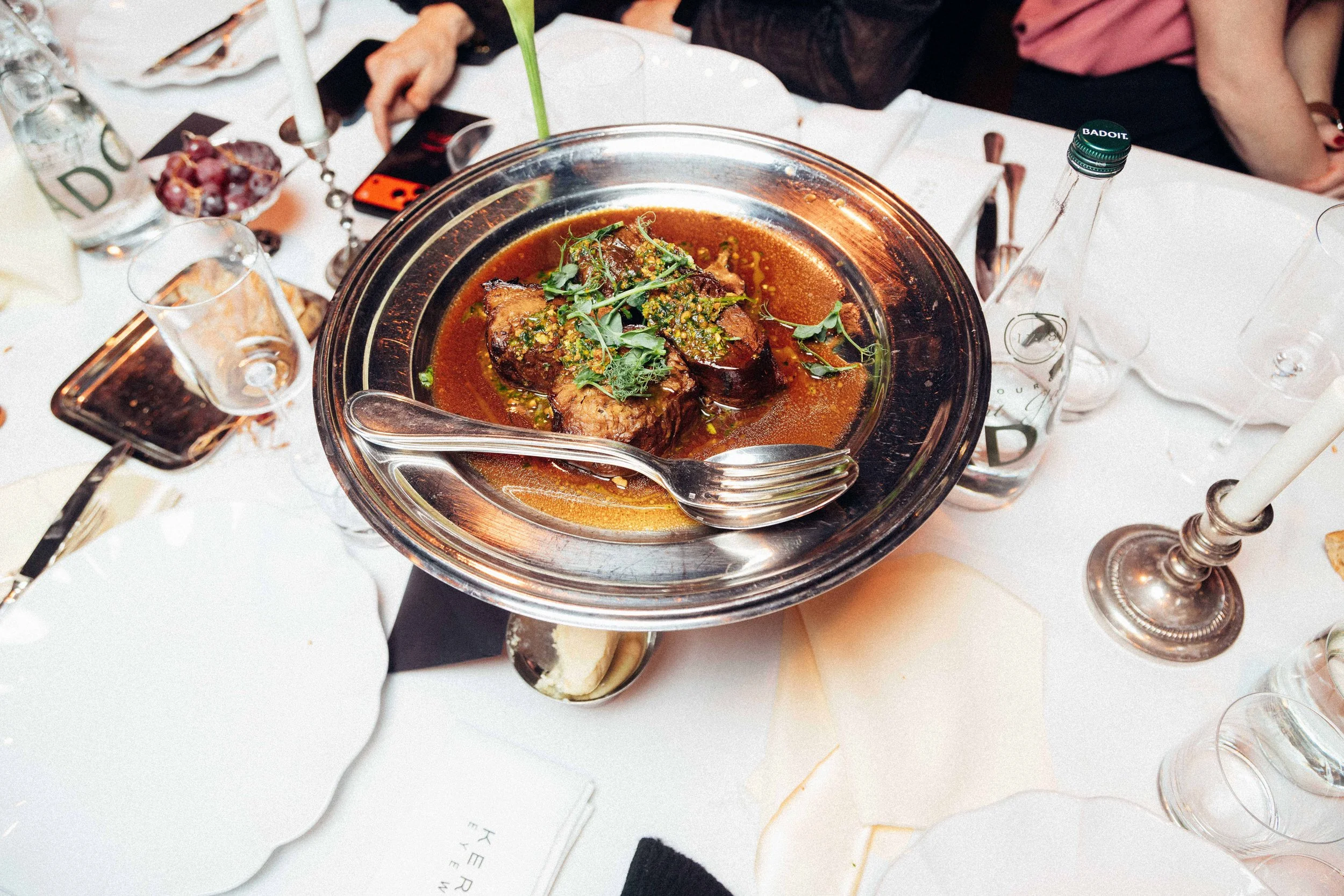 A plated dish with cooked meat in a dark sauce, garnished with herbs, on a metal serving tray on a dining table surrounded by glasses, bottles, and tableware.