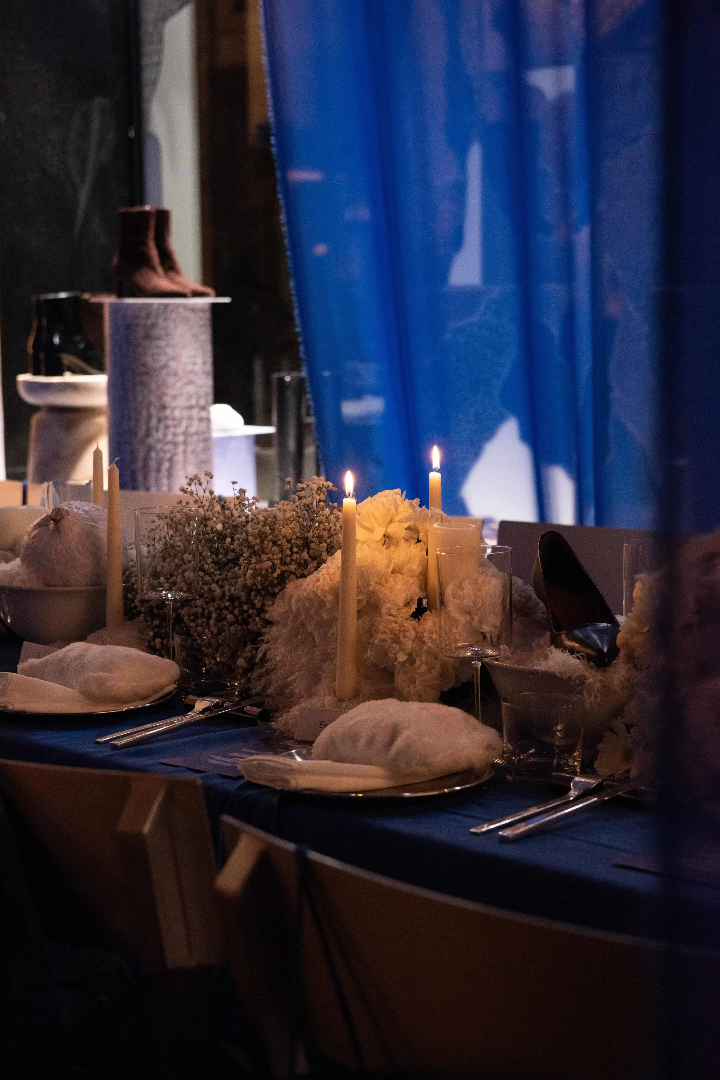 Elegant dining table with white flowers, candles, and place settings, dimly lit with blue curtains in background.