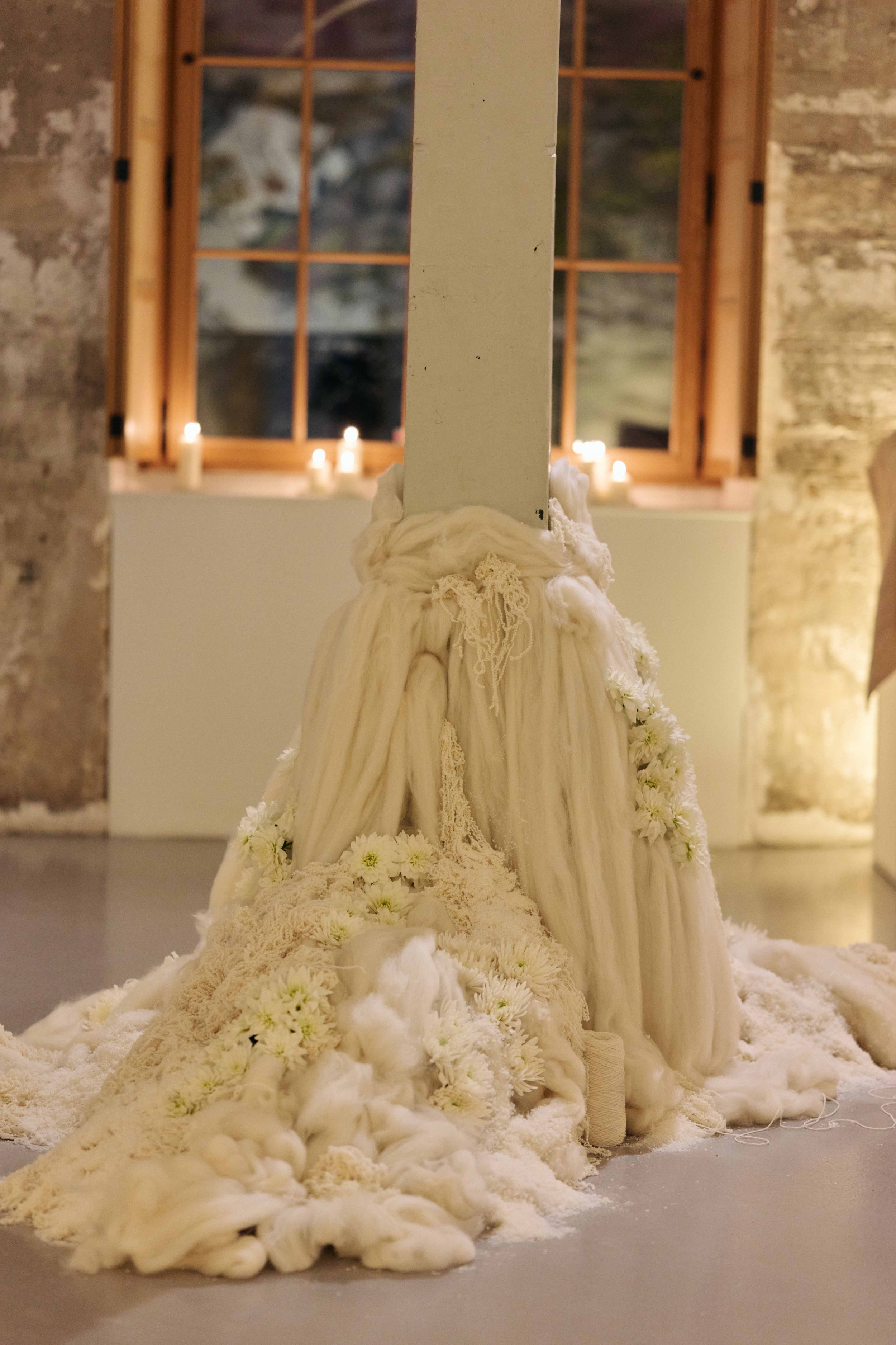 A white fabric draped around a vertical pole with white flowers and lace decorations, inside a room with stone walls, wooden window frames, and lit candles in the background.