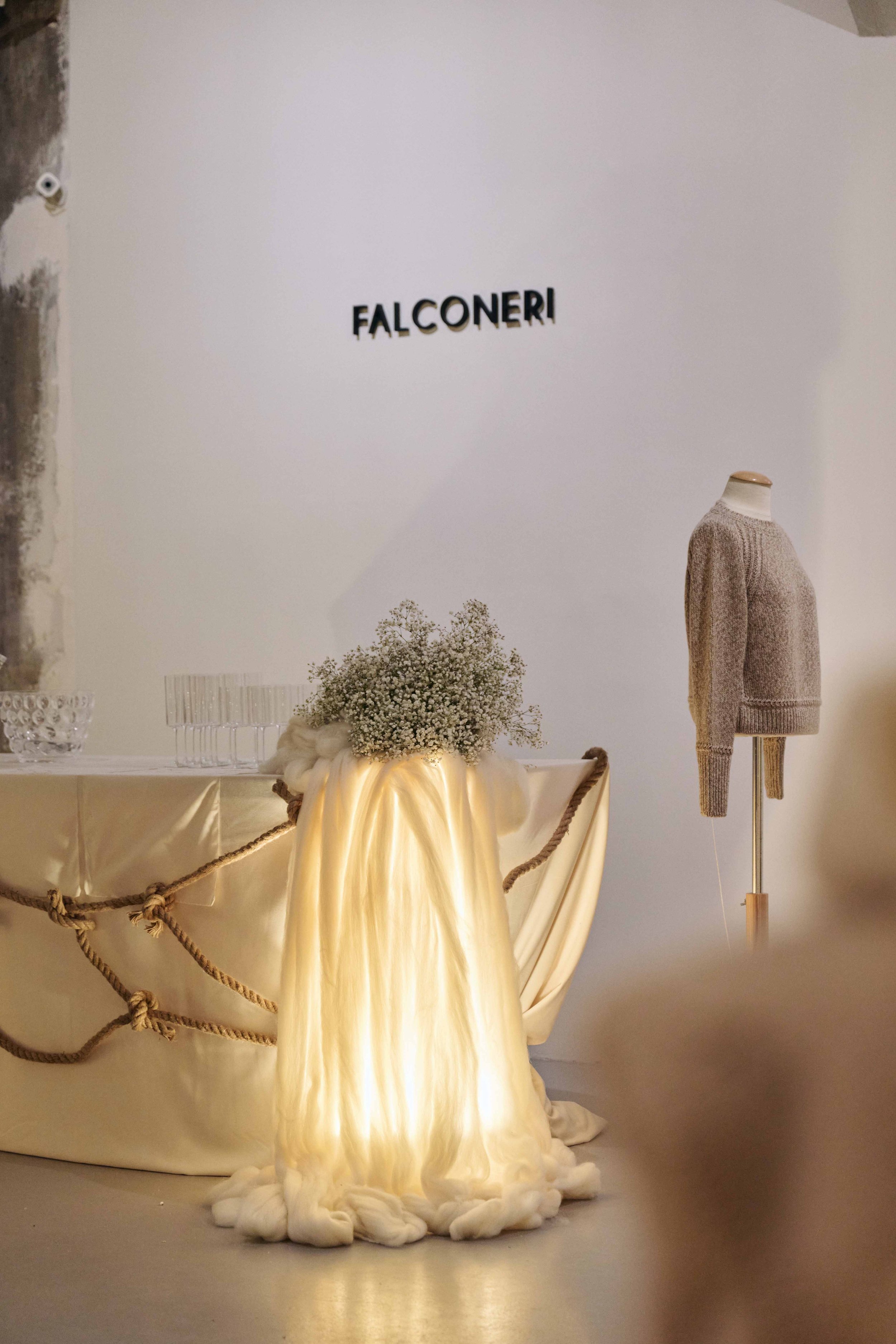 Display of a retail store named Falconeri with a glowing fabric centerpiece, a bouquet of baby's breath flowers, a table with glasses, and a mannequin dressed in a neutral-colored sweater.