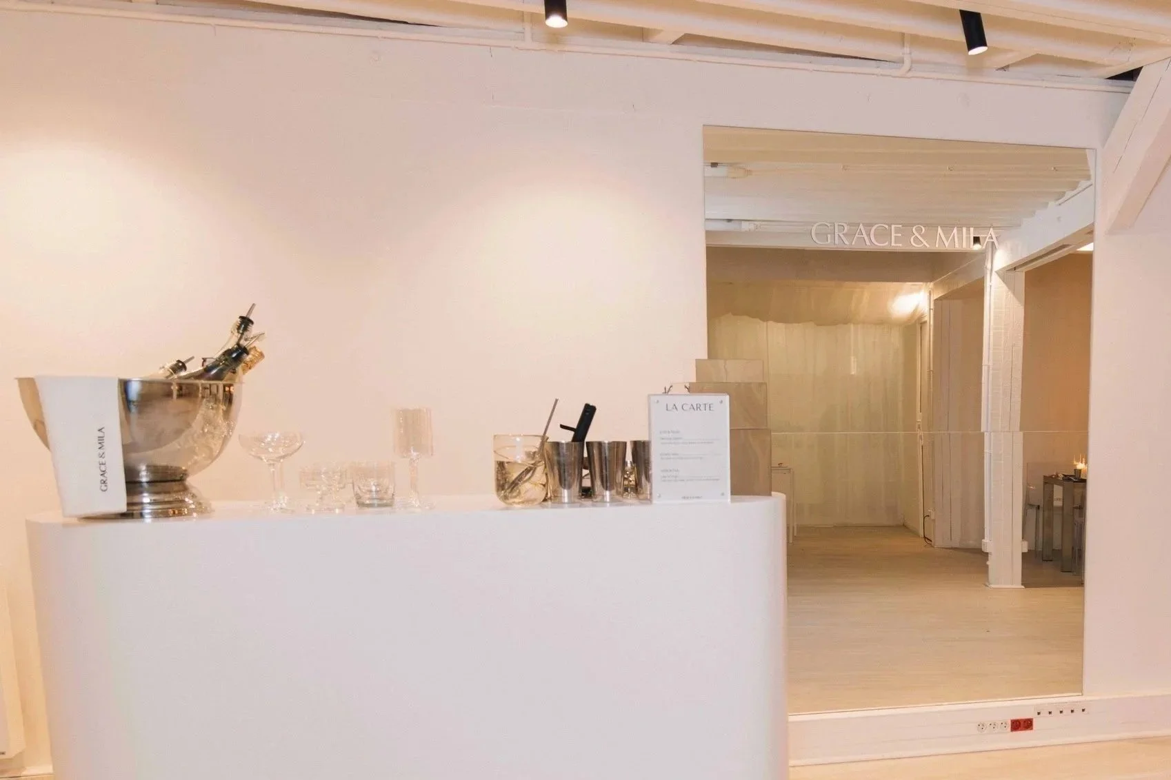 Interior of a modern, minimalist event space with white walls and light wood accents, including a table with champagne bottles, glasses, and a menu, and a nearby sign with the names 'GRACE & MILA'.