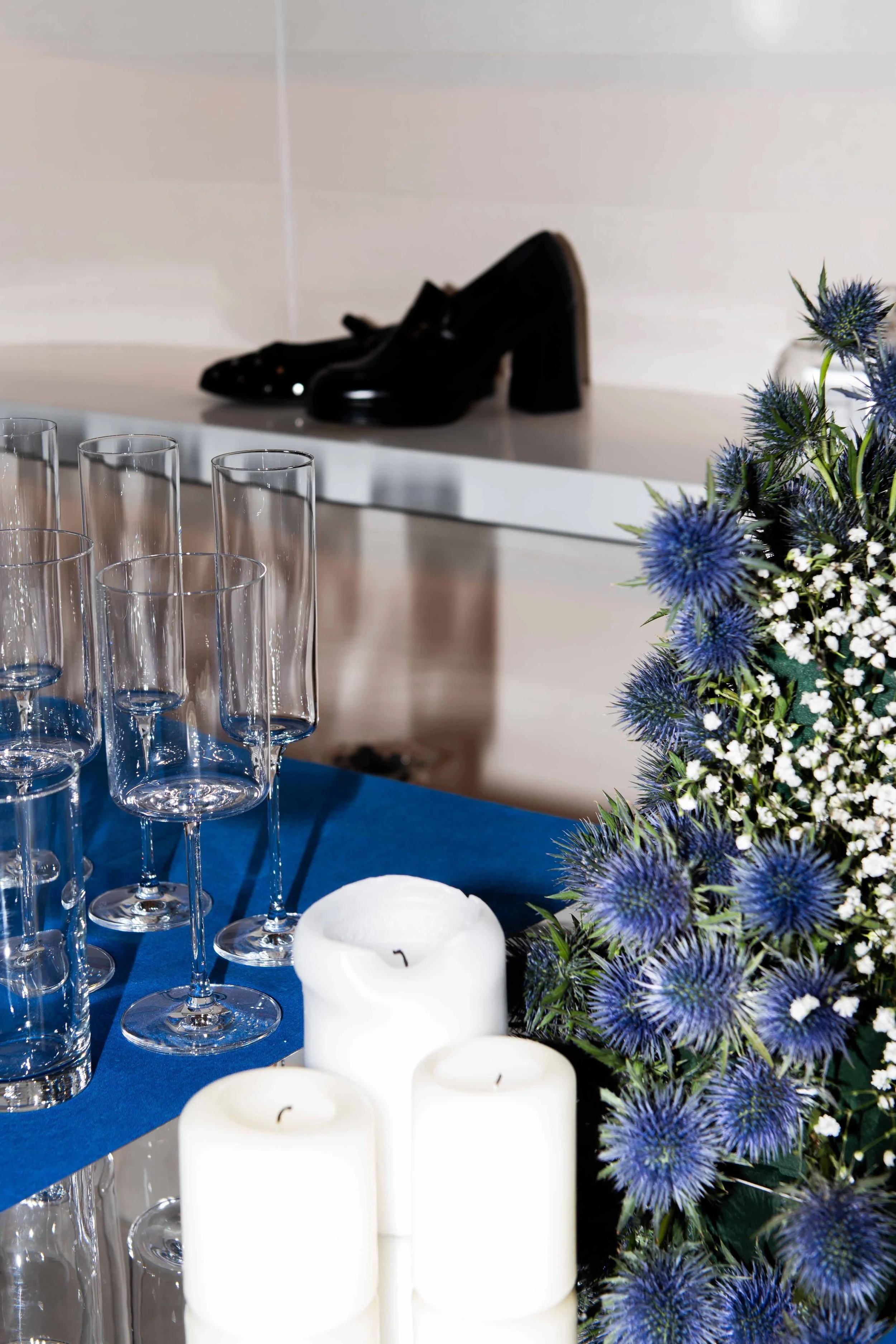 Black high-heeled shoes on a shelf, clear champagne flutes on a table with blue cloth, white candles, and a bouquet of blue thistle flowers and small white filler flowers.