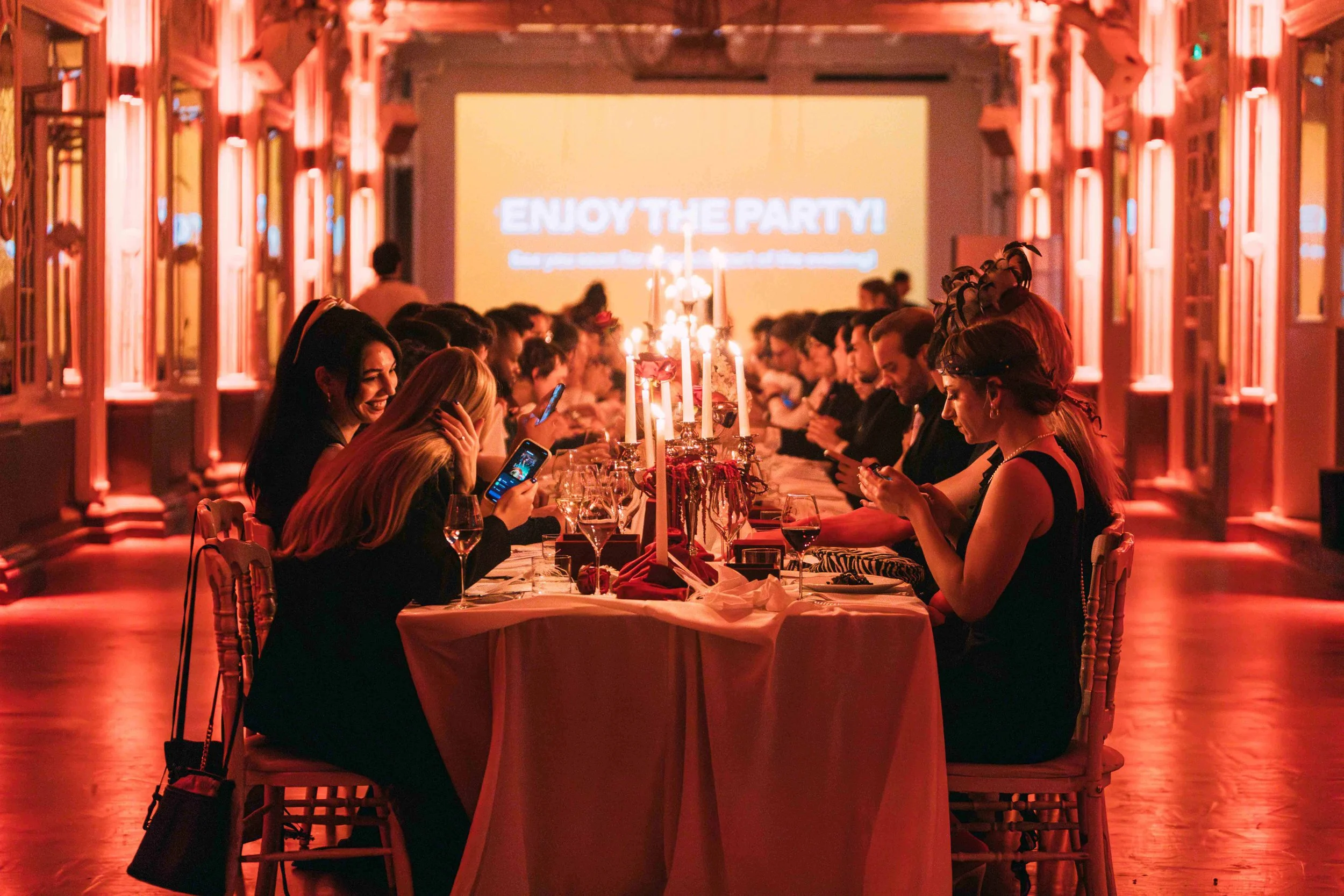 People sitting around a long banquet table with candles, enjoying a party in a dimly lit, ornate room, with a projection screen that says 'Enjoy the Party!' in the background.