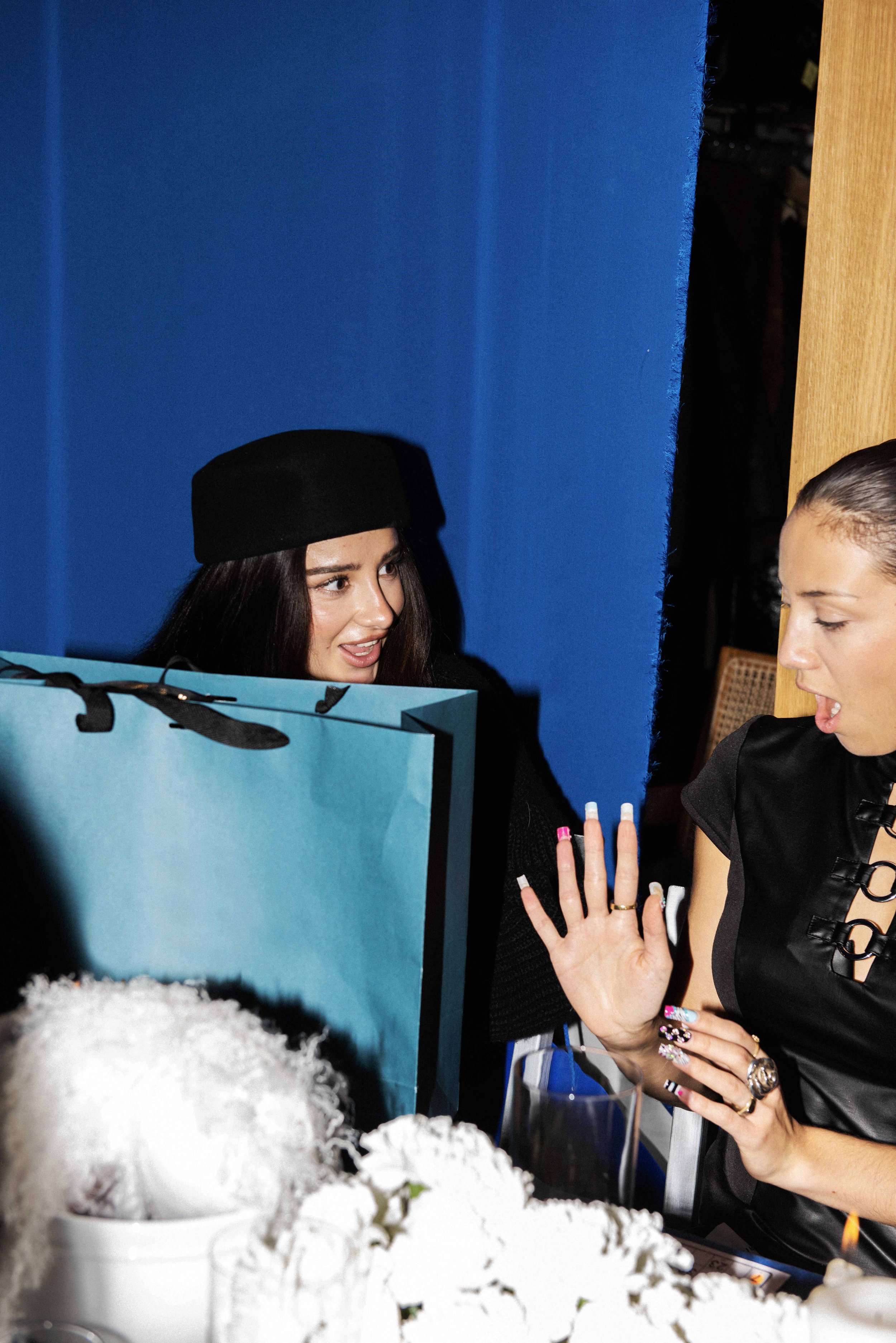 Two women engaged in conversation at a social gathering, one with long dark hair wearing a black beret, and the other with light hair, holding a hand up with colorful nails, amidst floral decorations and a large shopping bag.