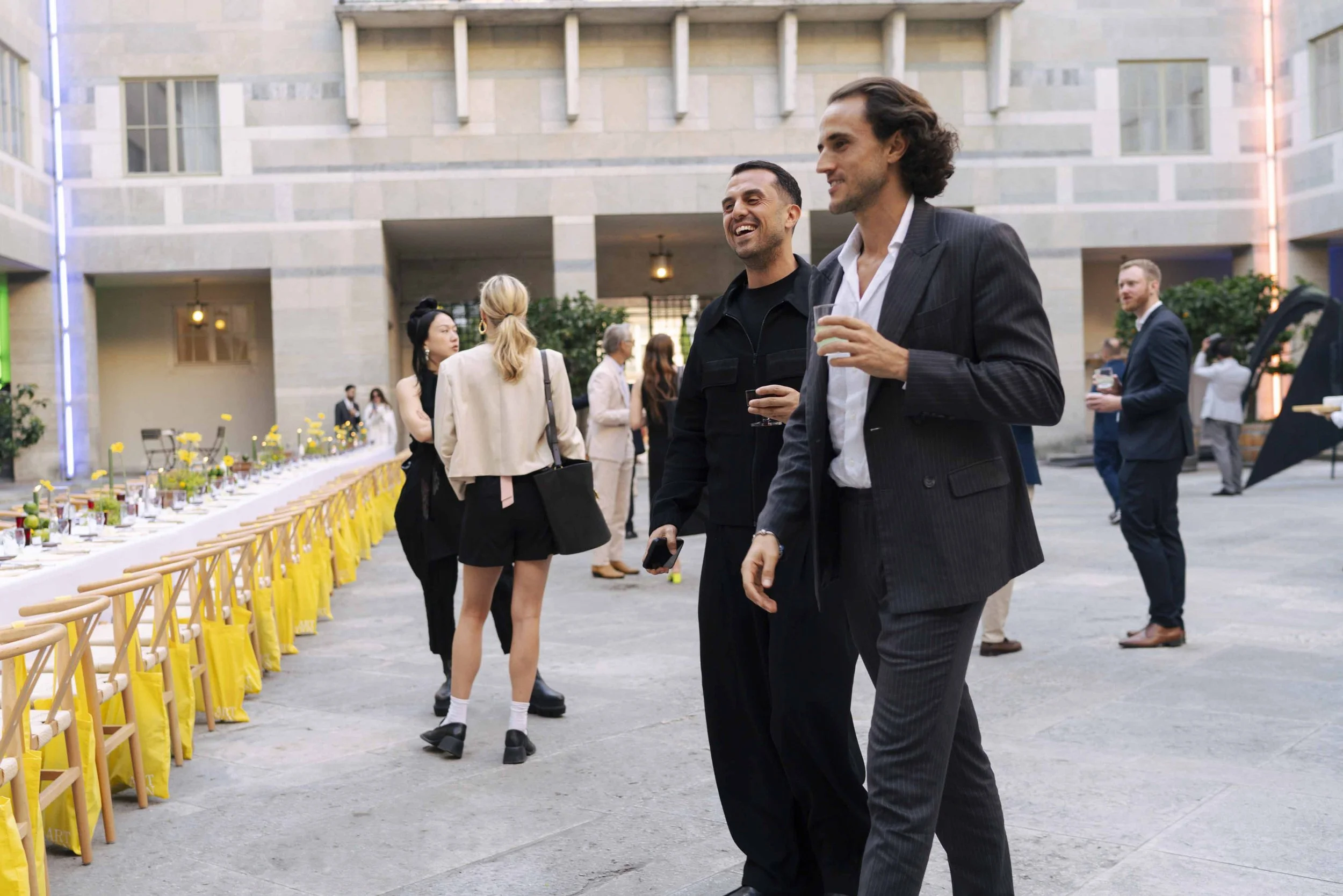 People socializing at an outdoor event in a courtyard with a long decorated table. Two men in the foreground are holding drinks and smiling, dressed in black and business suits.