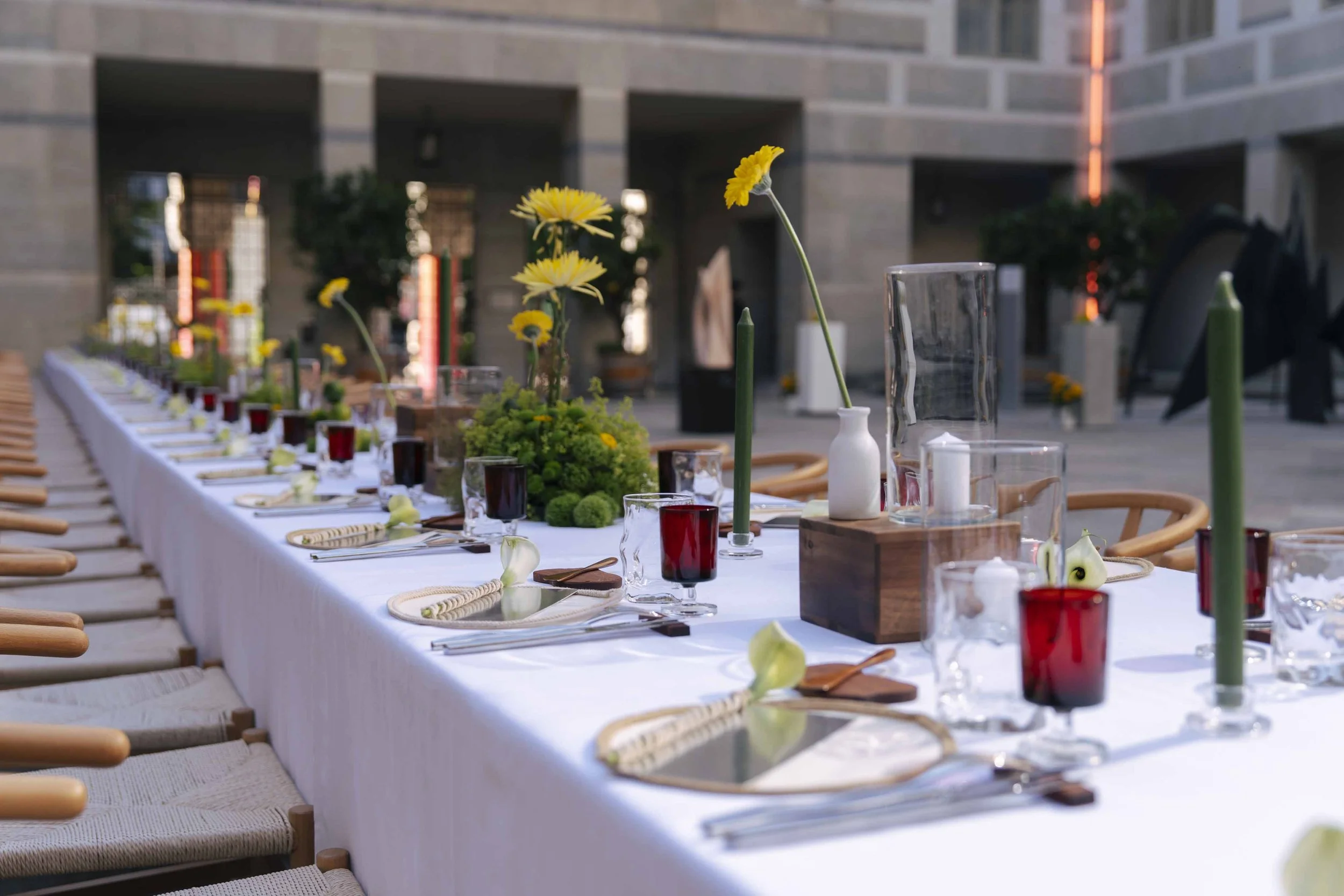 Long outdoor dining table decorated with flowers and candles in a modern building courtyard.