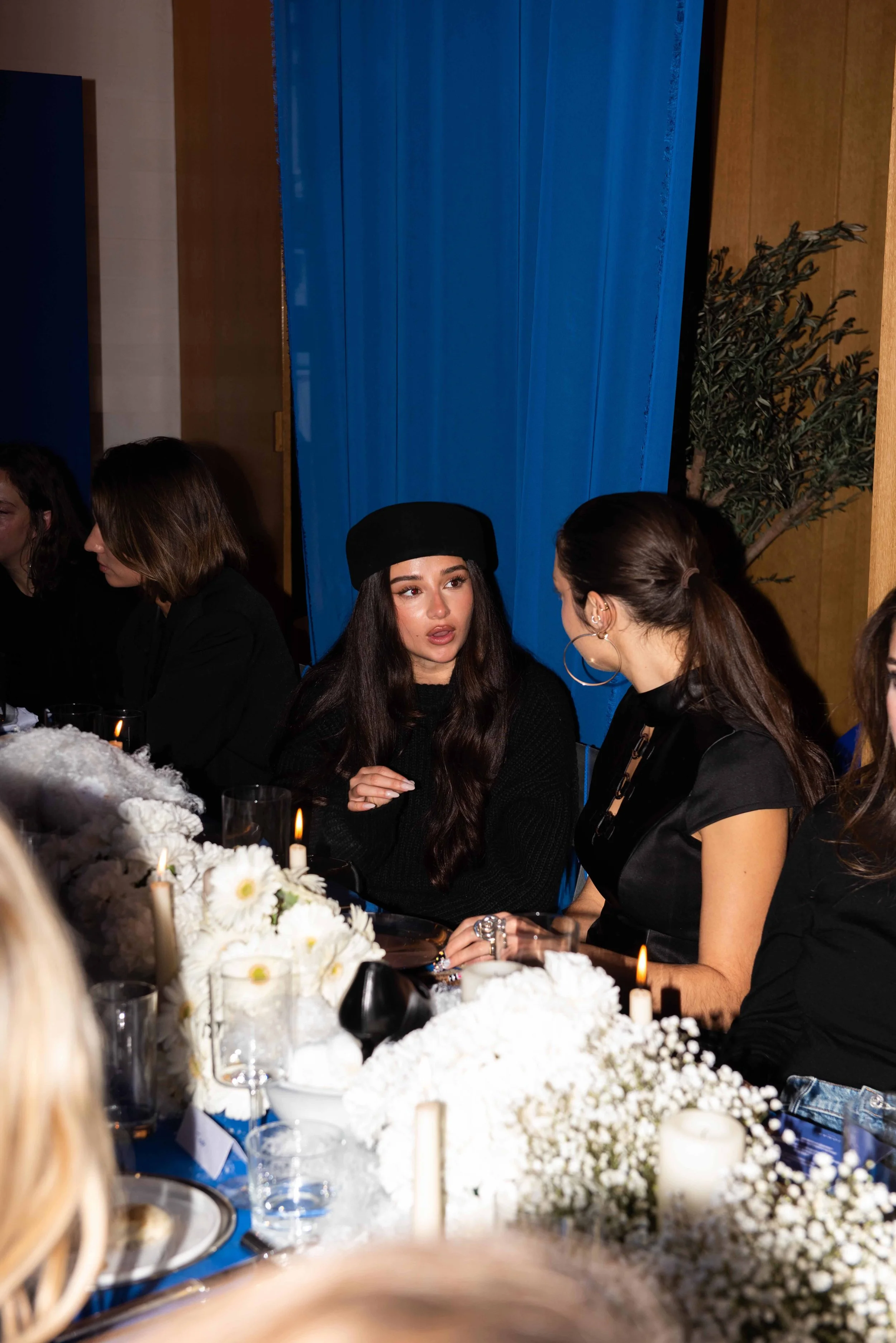 Two women engaged in conversation at a dinner party, with floral centerpieces and candles on the table, and a blue curtain in the background.