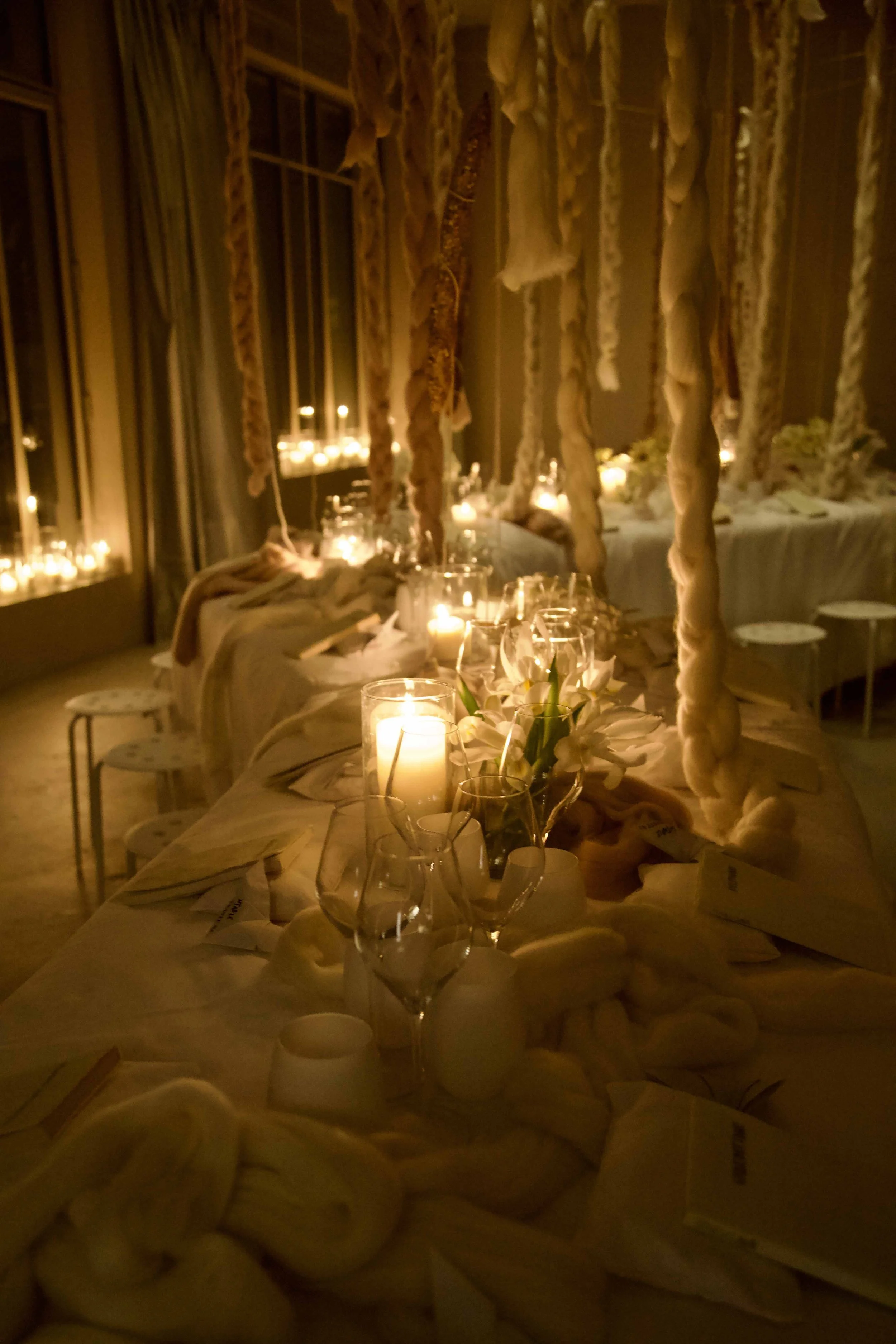 A long dining table decorated with candles, white flowers, and soft fabric, set for an elegant event in a warmly lit room with hanging yarn or fabric decorations and candlelit windows.
