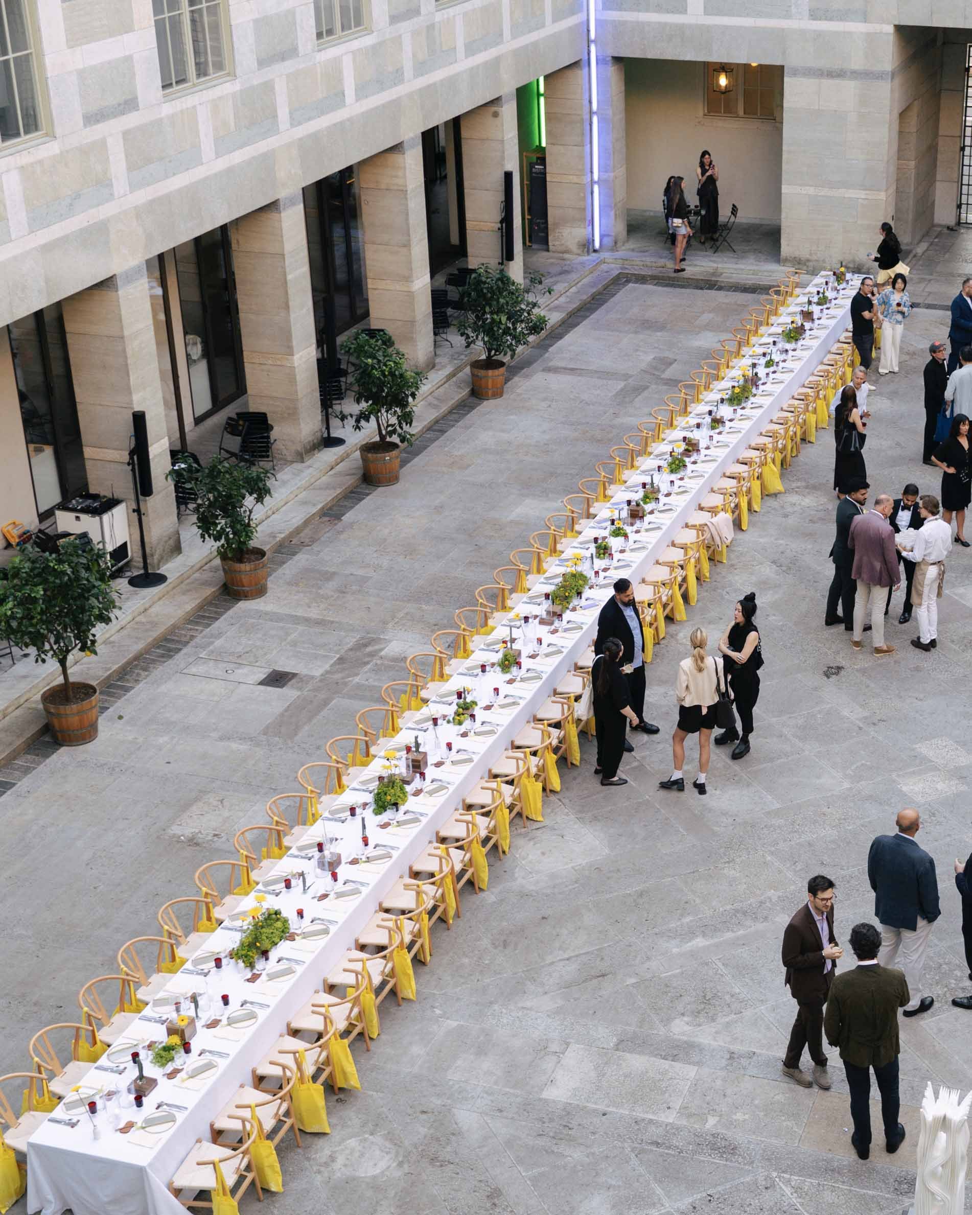 Long banquet table set up for a formal event in an open indoor courtyard with people mingling and chatting.