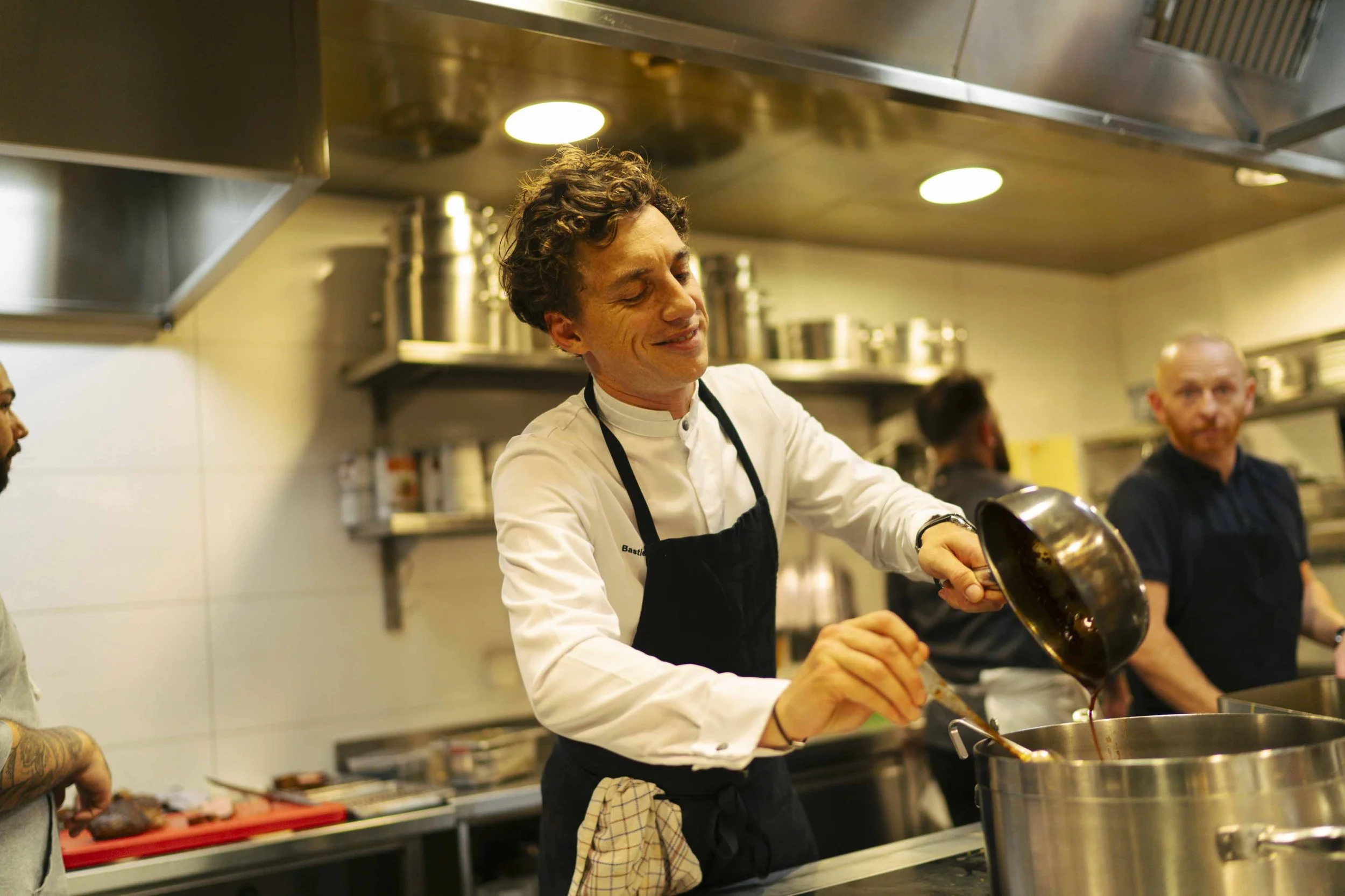 Chef pouring sauce into a pot in a professional kitchen, with other chefs working in the background.