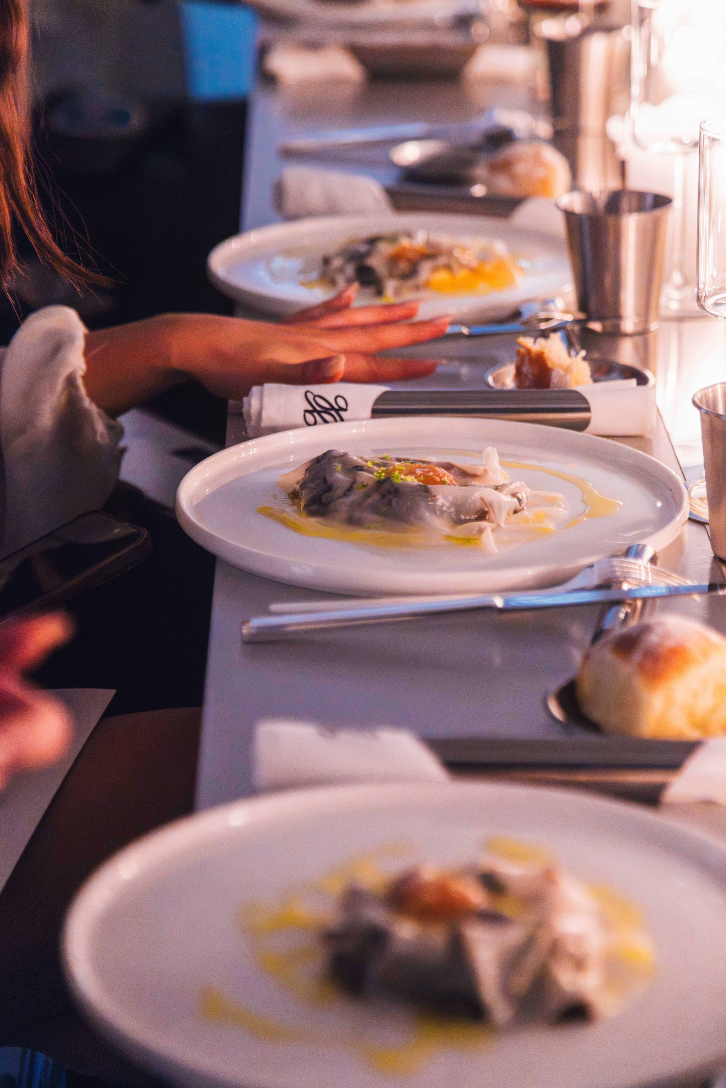 Several white plates with raw oysters topped with sauce, garnished with herbs, on a dining table set with utensils and bread rolls, with a person reaching for food.