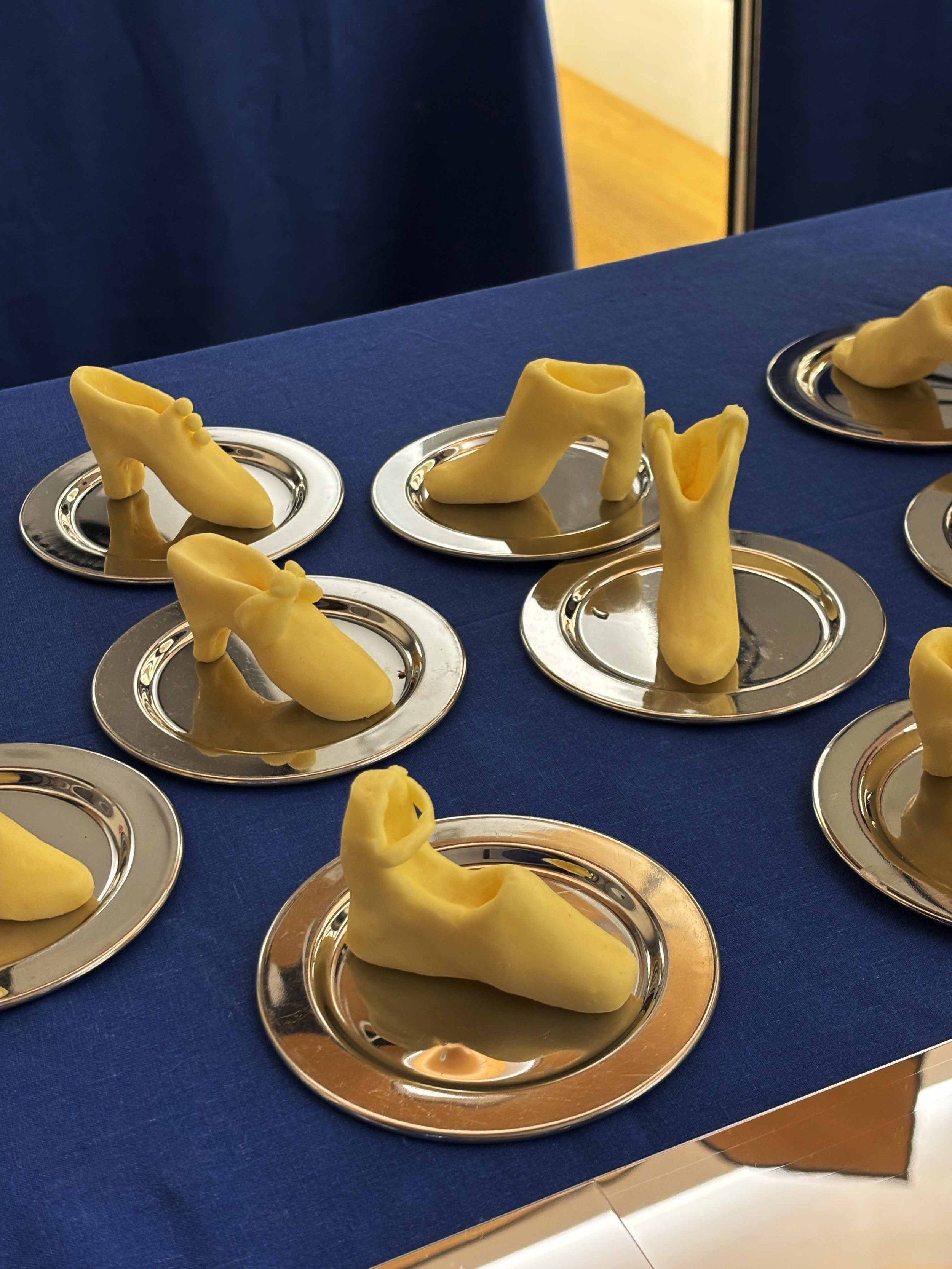 Several yellow fondant shoes placed on small silver trays on a blue tablecloth.